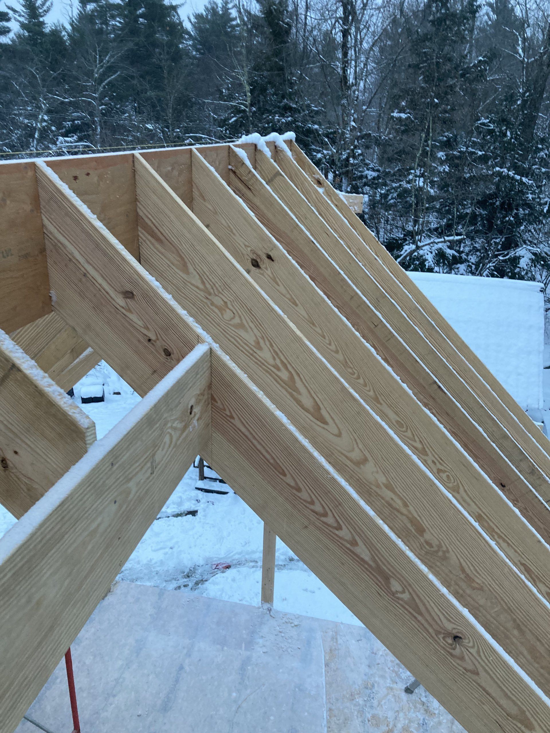 Wooden roof trusses under construction in a snowy outdoor setting, with trees in the background