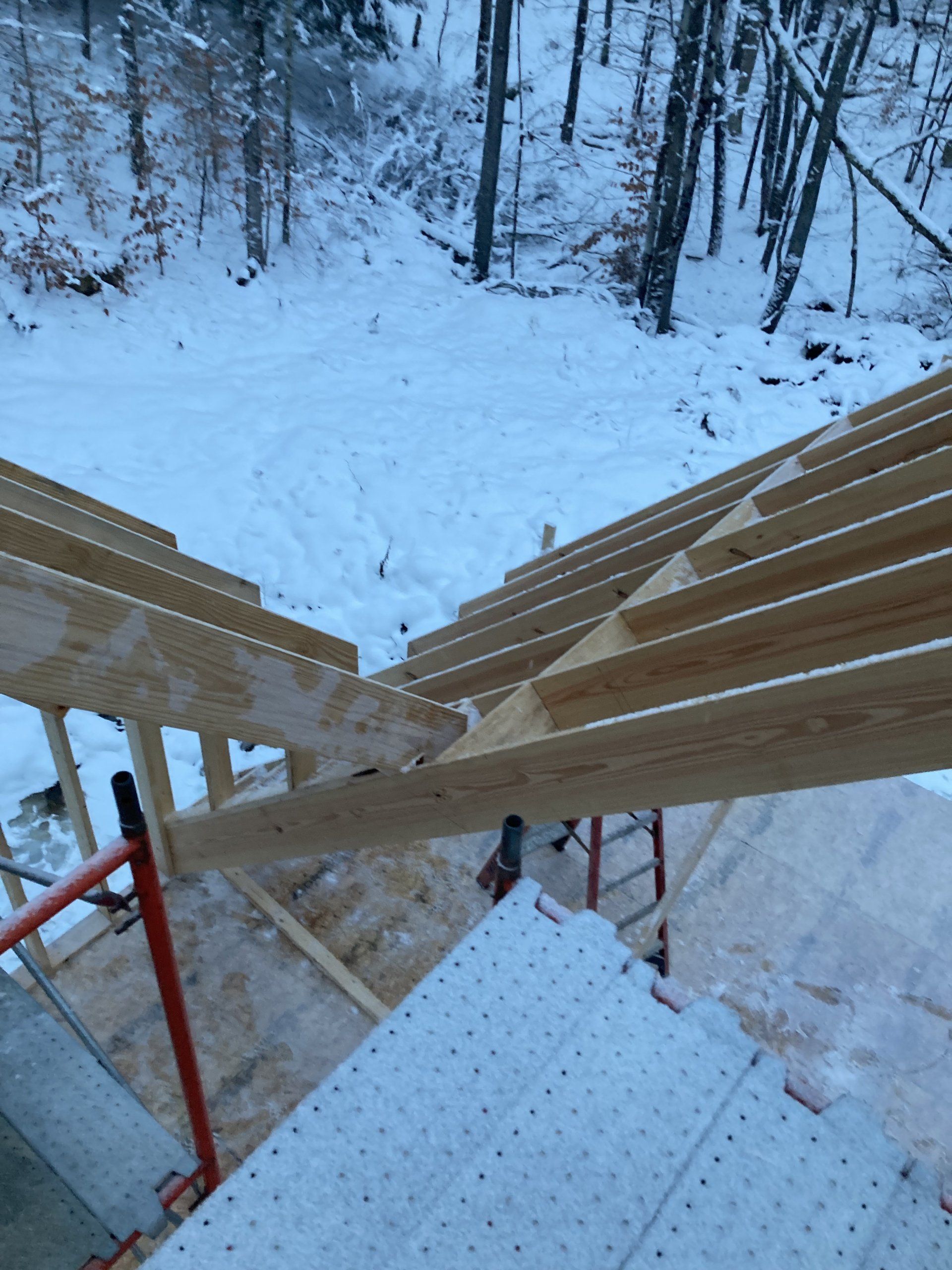 Snowy outdoor worksite seen from a scaffold with wooden planks and a red ladder in the foreground.