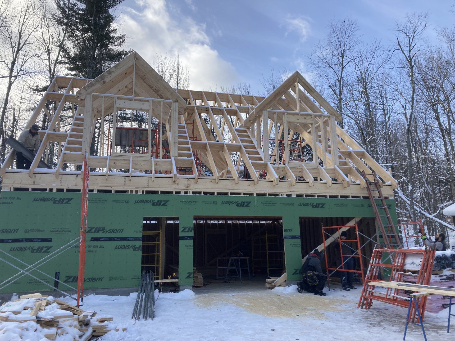 House under construction with exposed roof framing and green sheathing in a snowy wooded area
