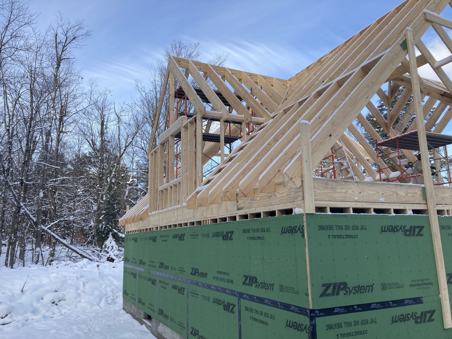 Partially built house with wooden roof trusses on a green foundation in a snowy wooded area