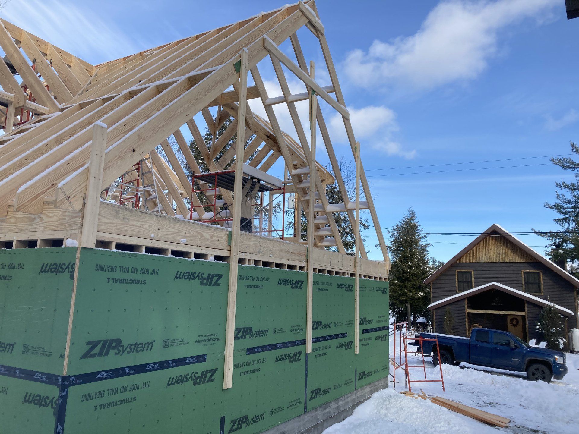 House under construction with exposed wooden roof framing beside a snowy driveway and garage.