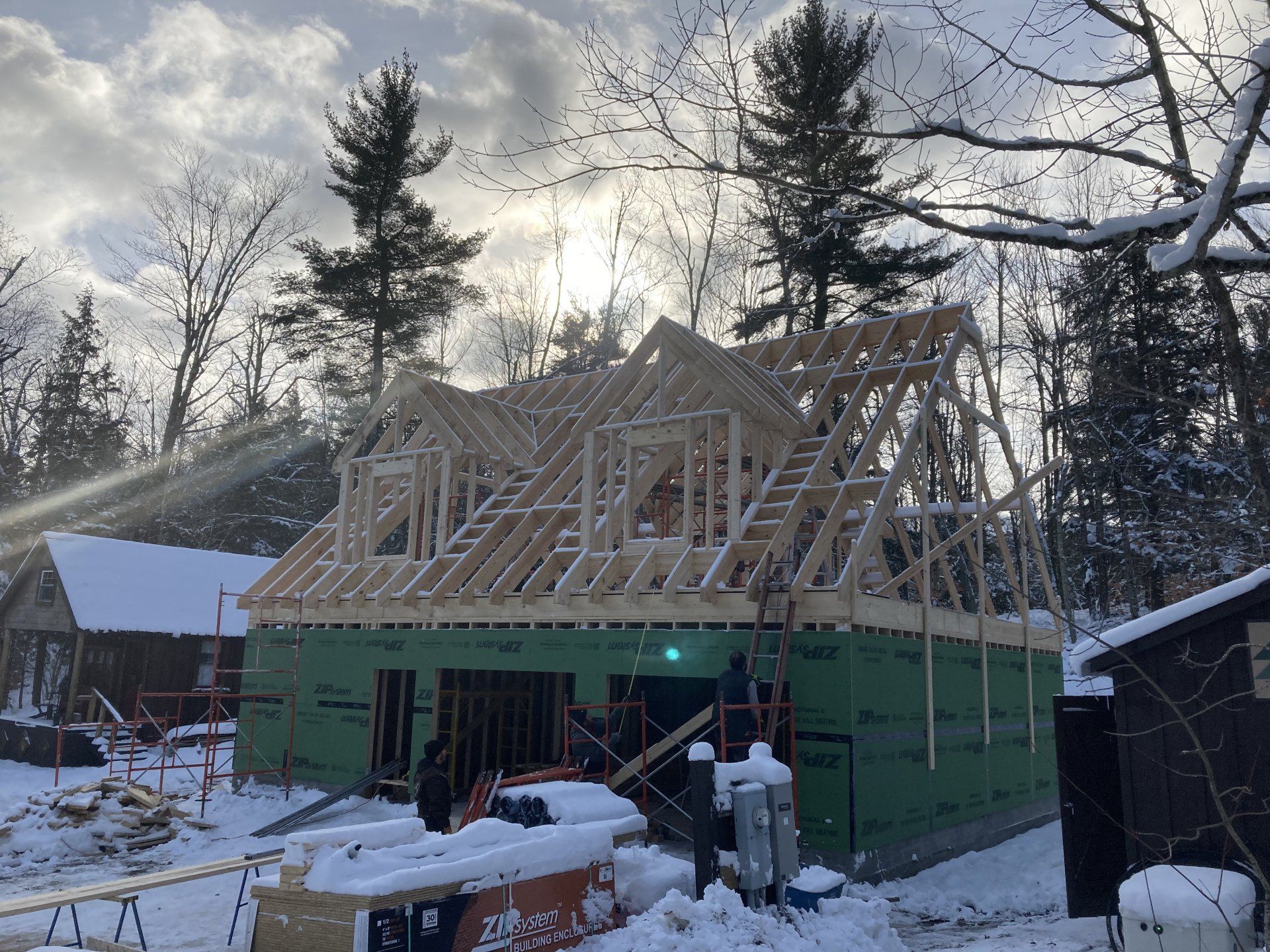Snow-covered house under construction with exposed roof framing and green exterior wrap at dusk