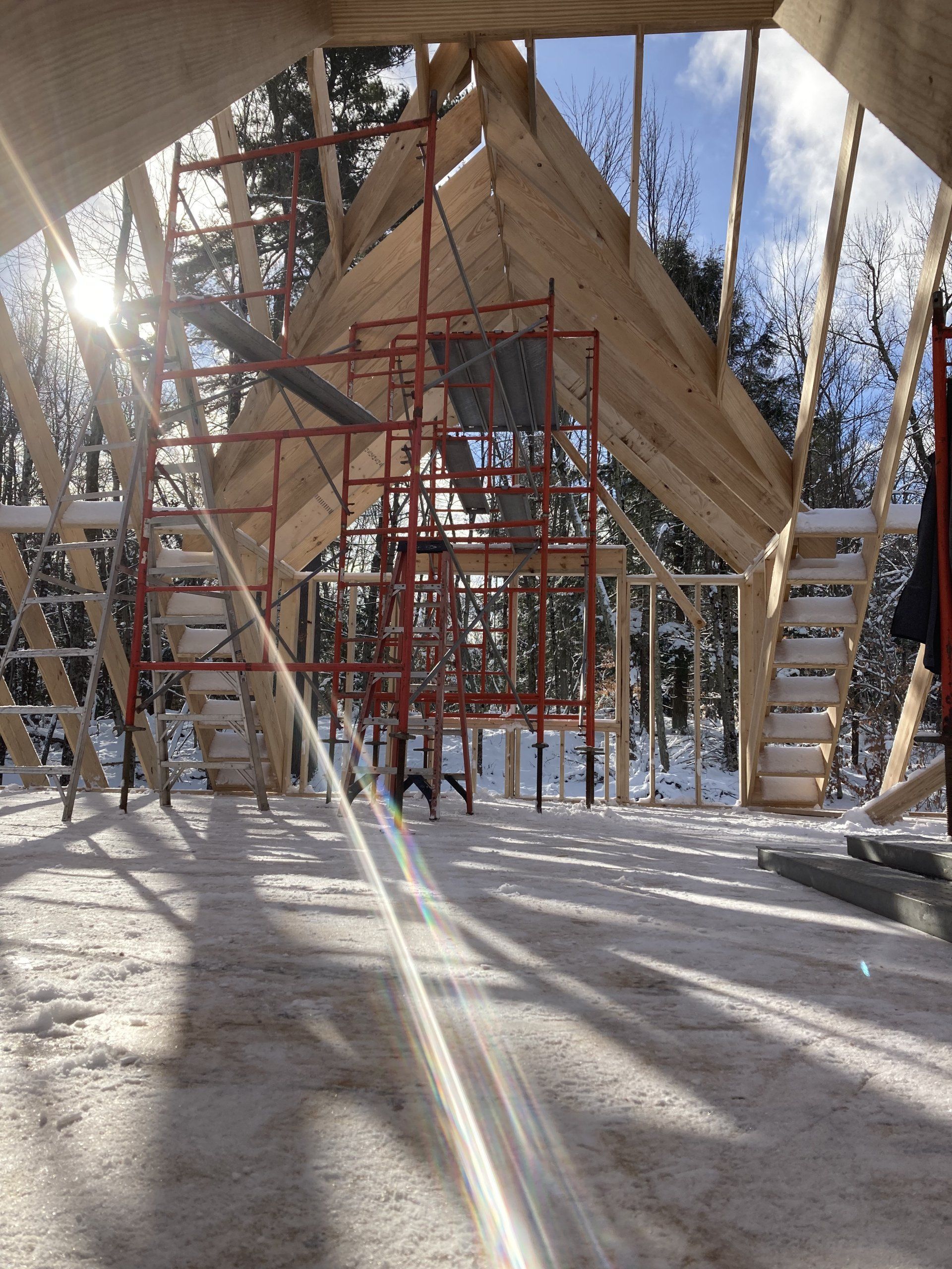 Wood-framed A-frame cabin under construction in a snowy forest, with red scaffolding and sunlight streaming through.
