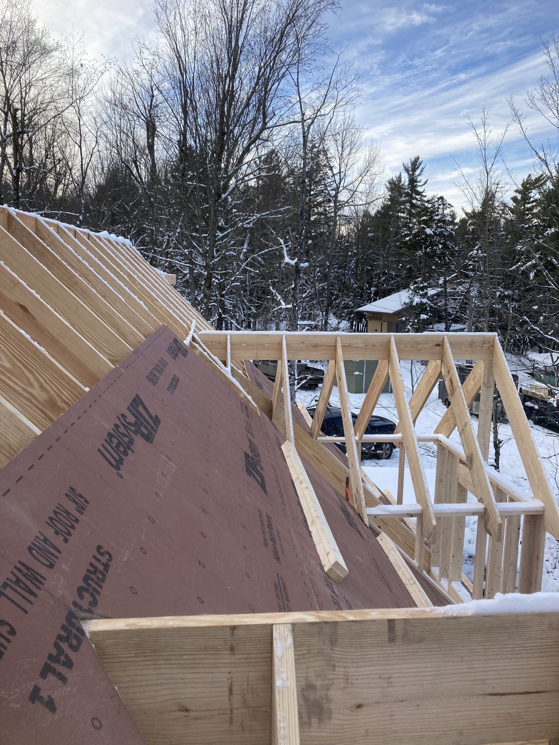 Wood-framed roof under construction with pink sheathing and snow on the ground under a cloudy sky
