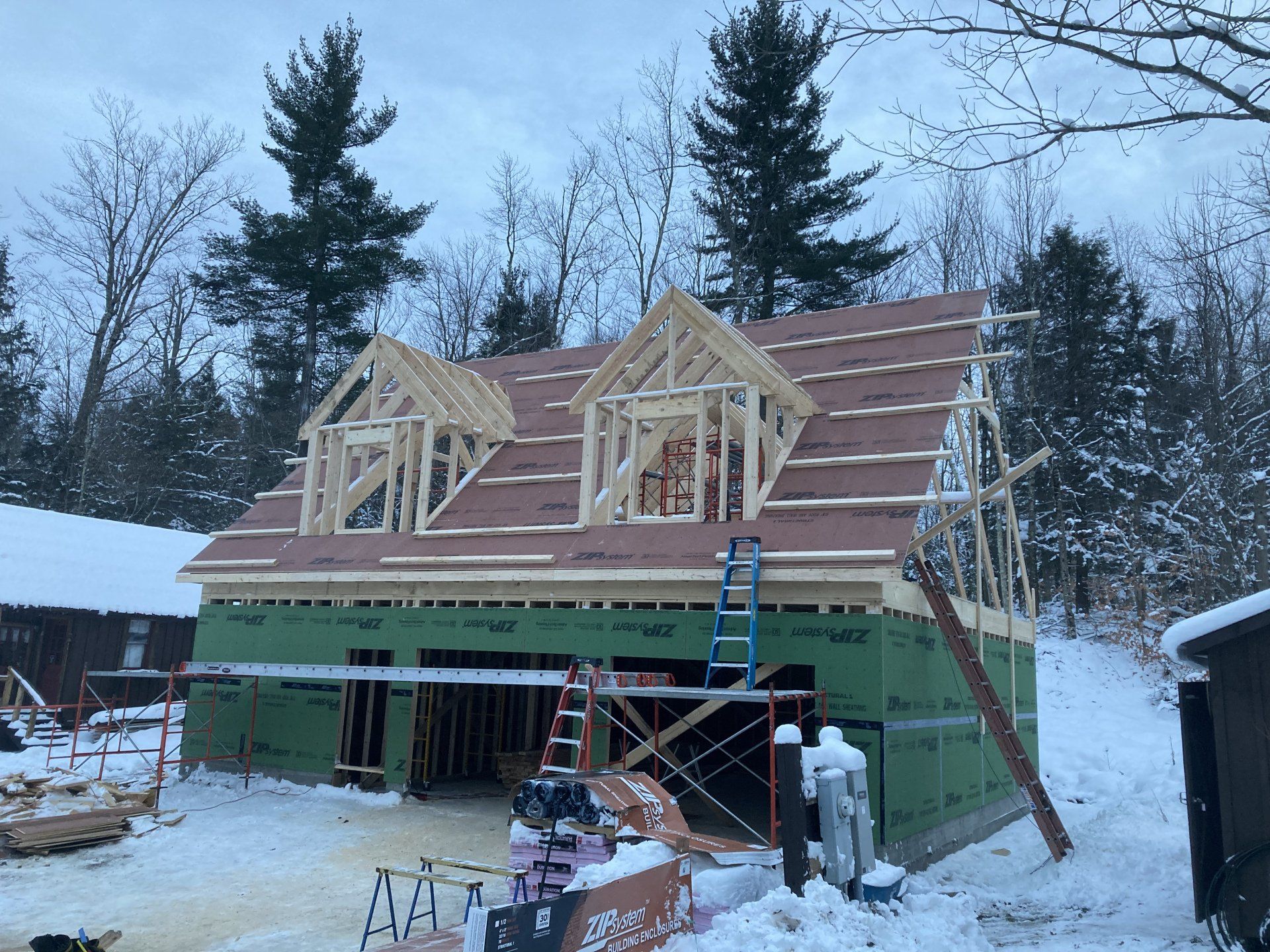 Snowy house under construction with roof framing, green siding, and ladders in a wooded yard