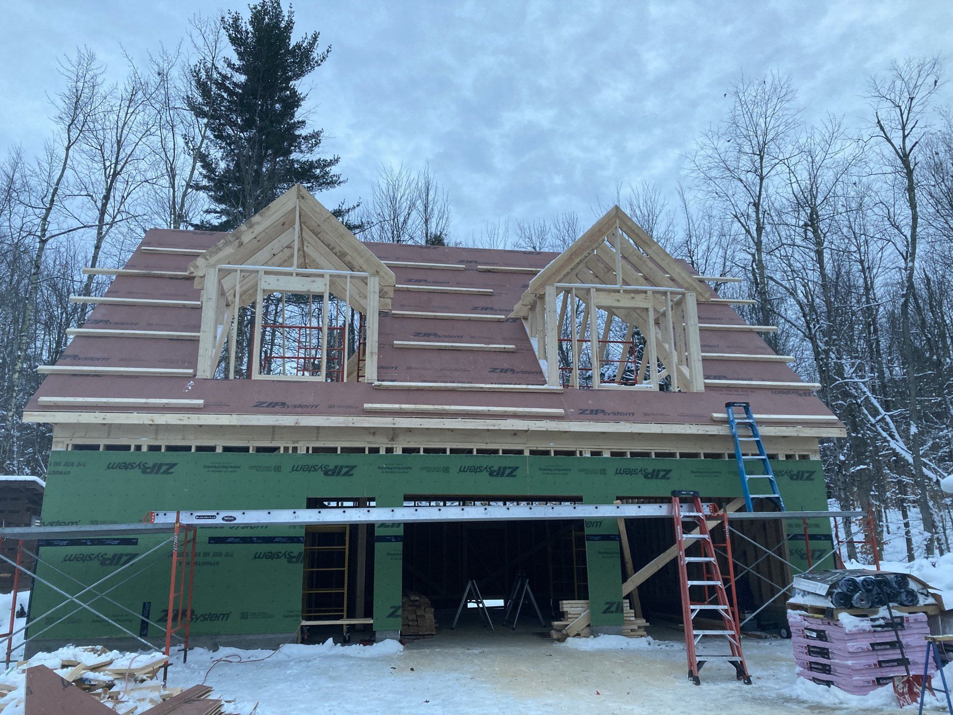 Two-story house under construction with green siding, unfinished roof, and scaffolding in a snowy wooded yard