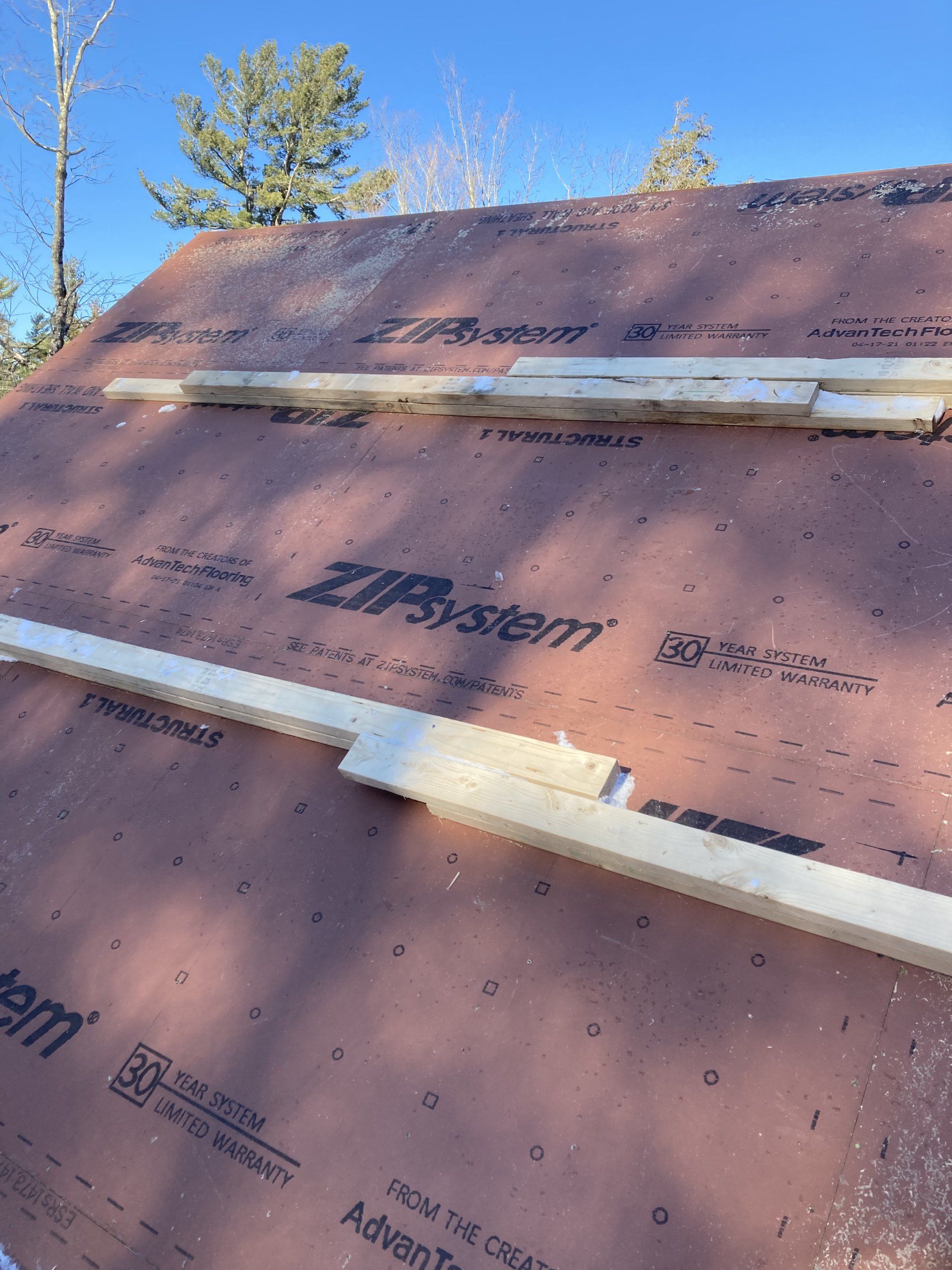 Pink roofing underlayment on a sloped roof with white trim strips and trees against a blue sky