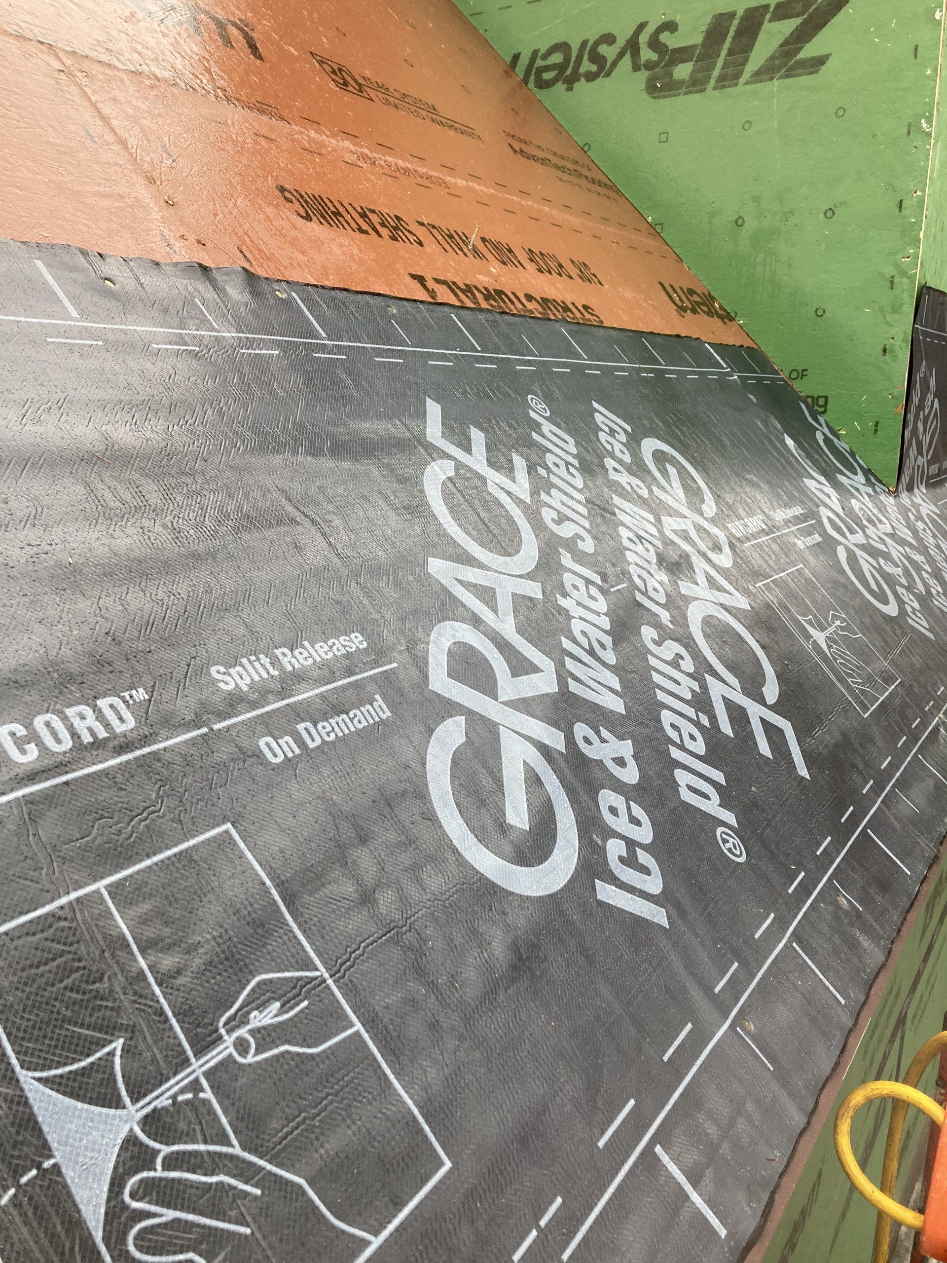 Close-up of printed roofing underlayment sheets in gray, orange, and green, with a yellow cable at right.