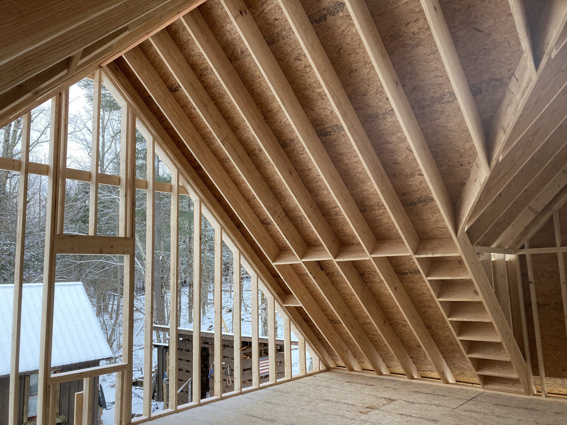 Exposed wooden roof framing inside an unfinished room with large windows and snowy trees outside