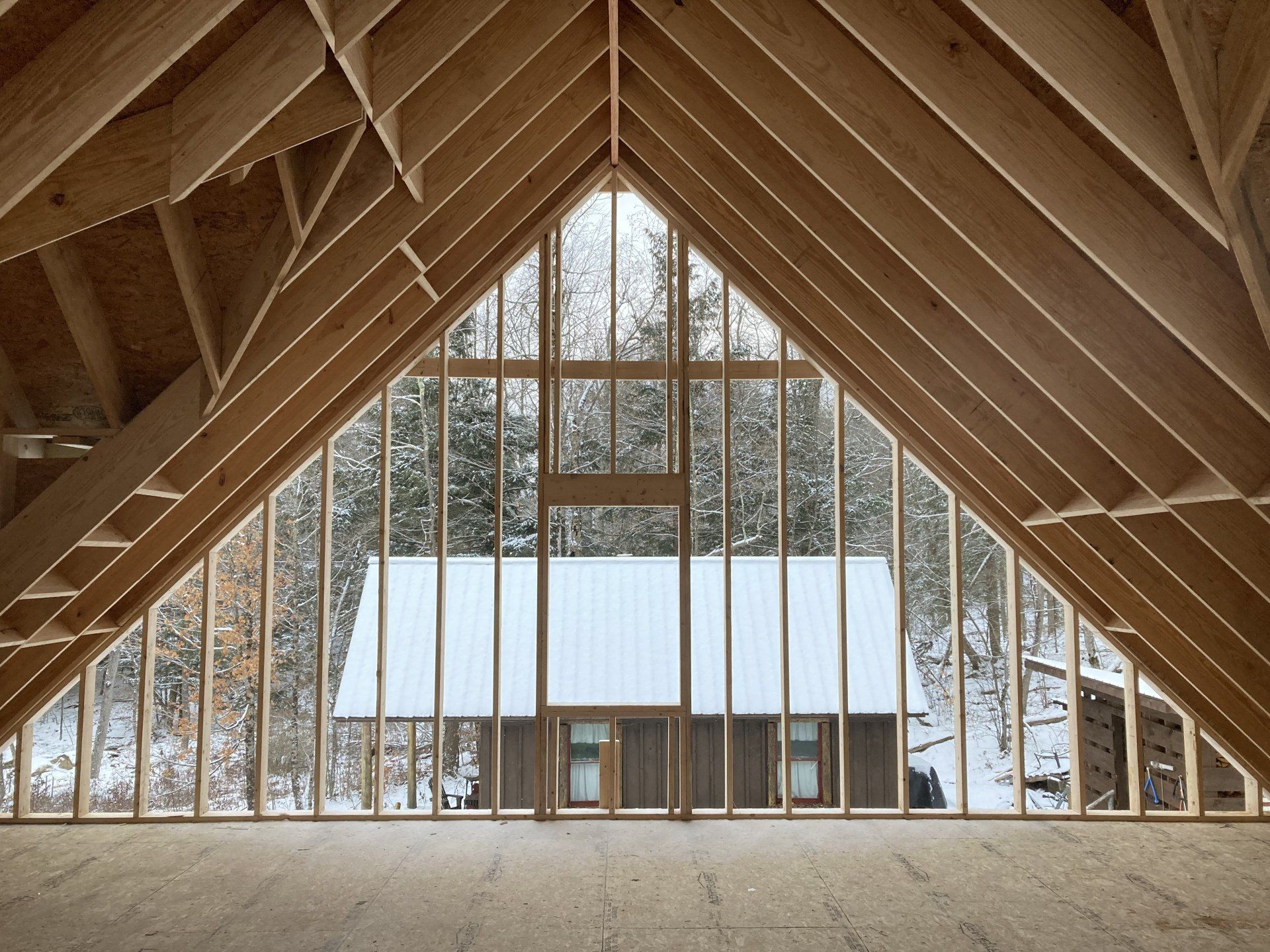 A wooden A-frame interior framing a snow-covered house and winter trees outside