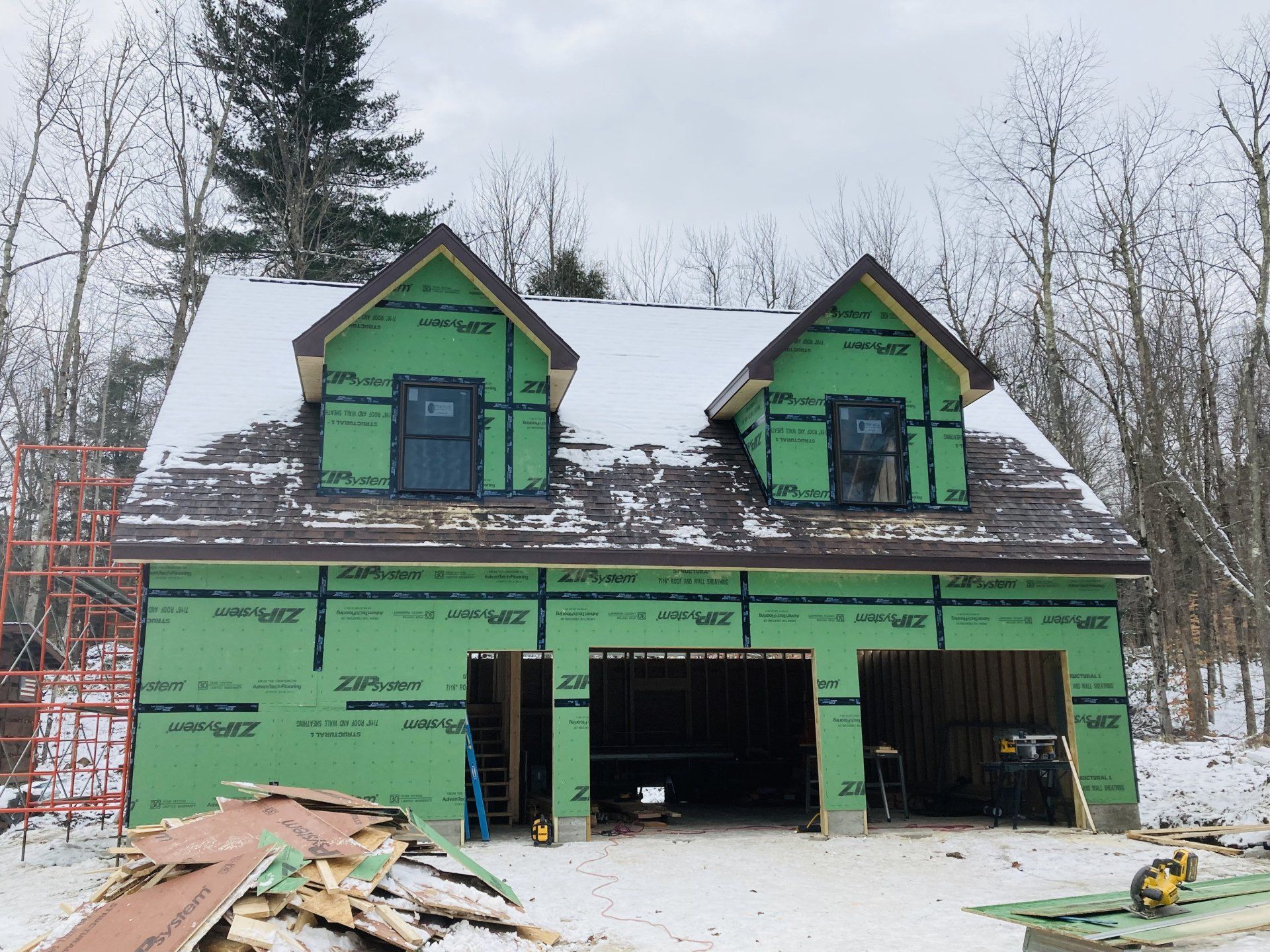 Green house under construction with two dormer windows and open garage bays, surrounded by snow.