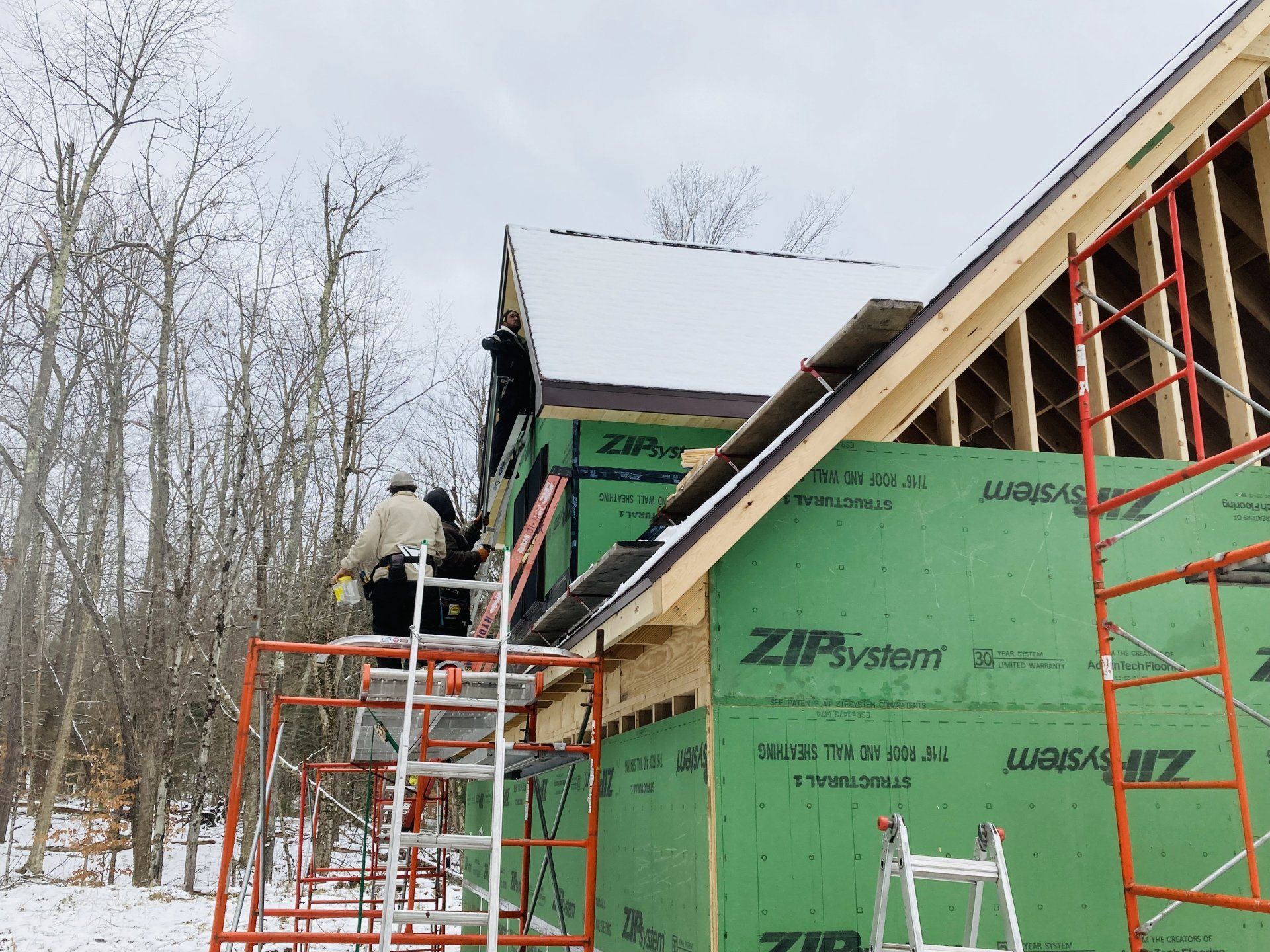 Workers on scaffolding install green sheathing on a house under construction in a snowy wooded area