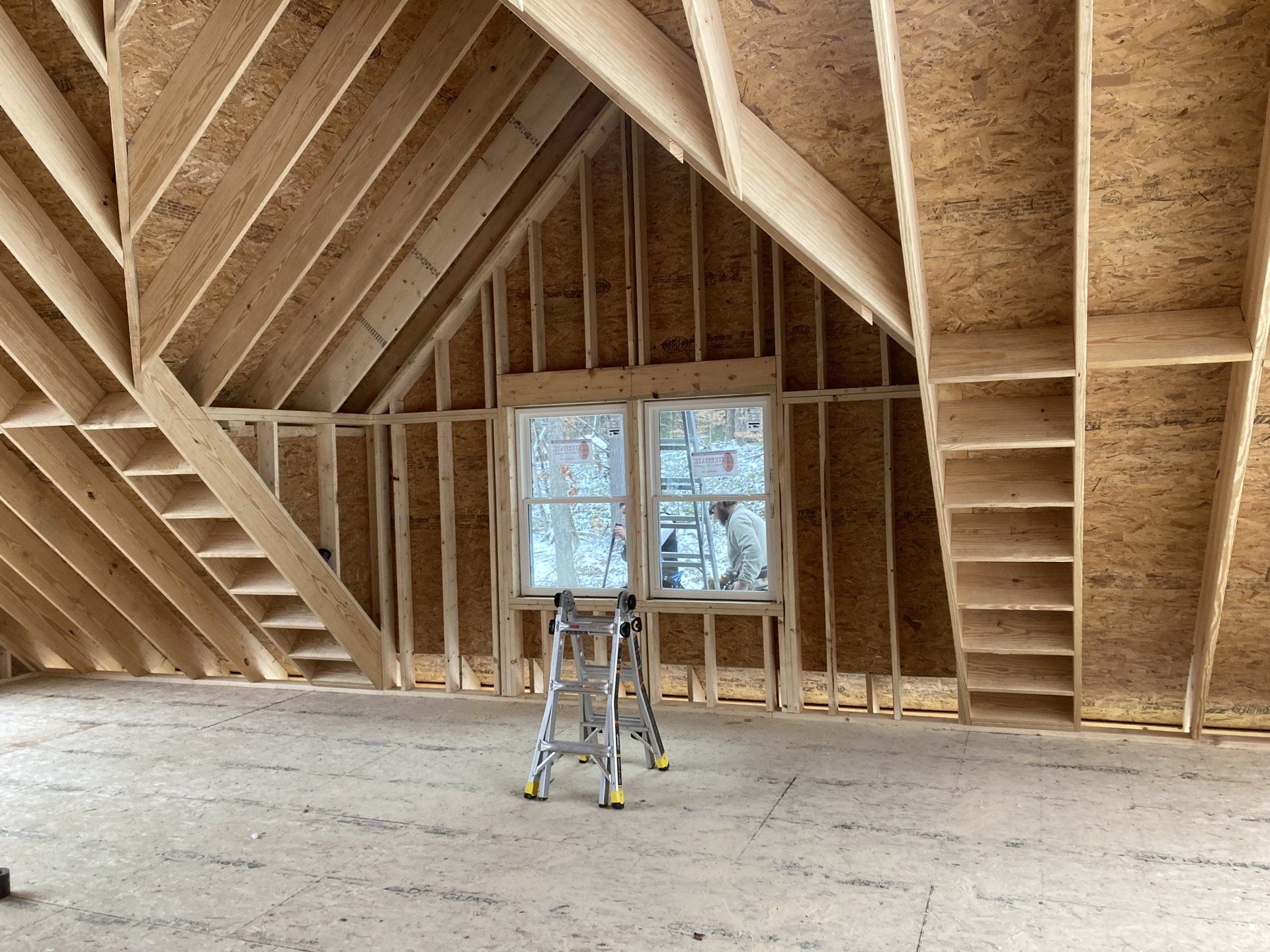Unfinished attic interior with exposed wooden framing, a small ladder, and a window at the far end.