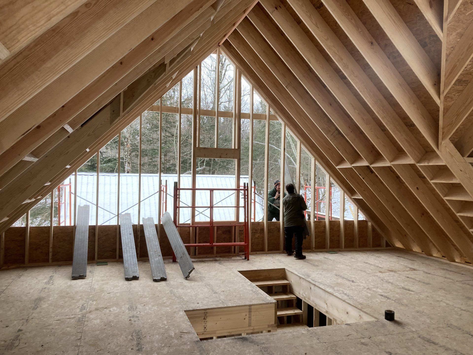 Wood-framed attic under construction with roof trusses, floor opening, and two workers near the window wall