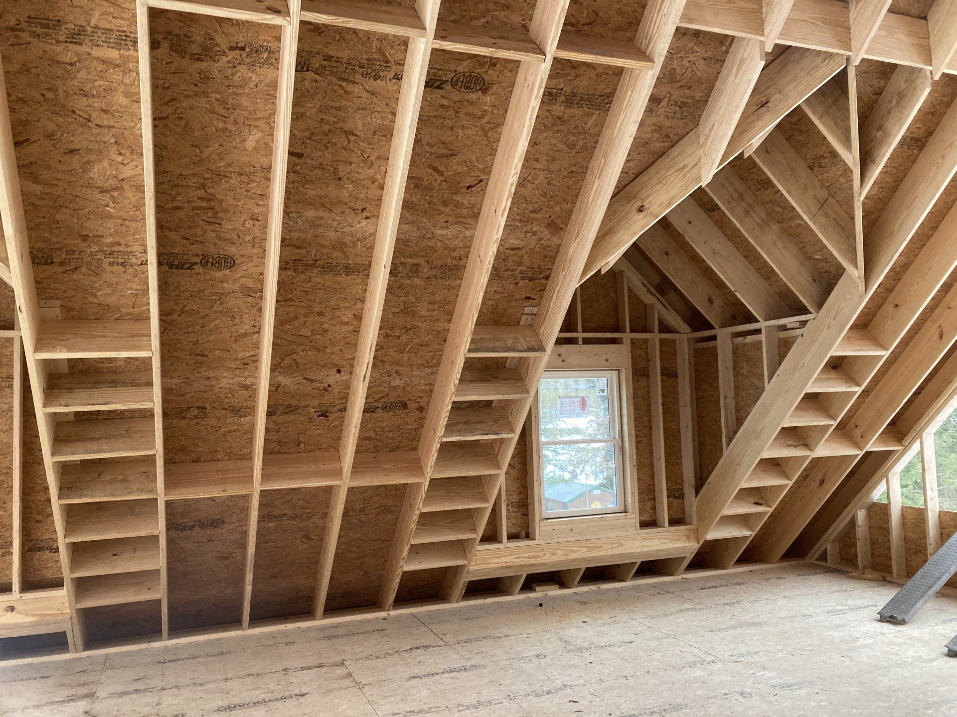 Unfinished attic framing with exposed wooden rafters, floor joists, and a small window.