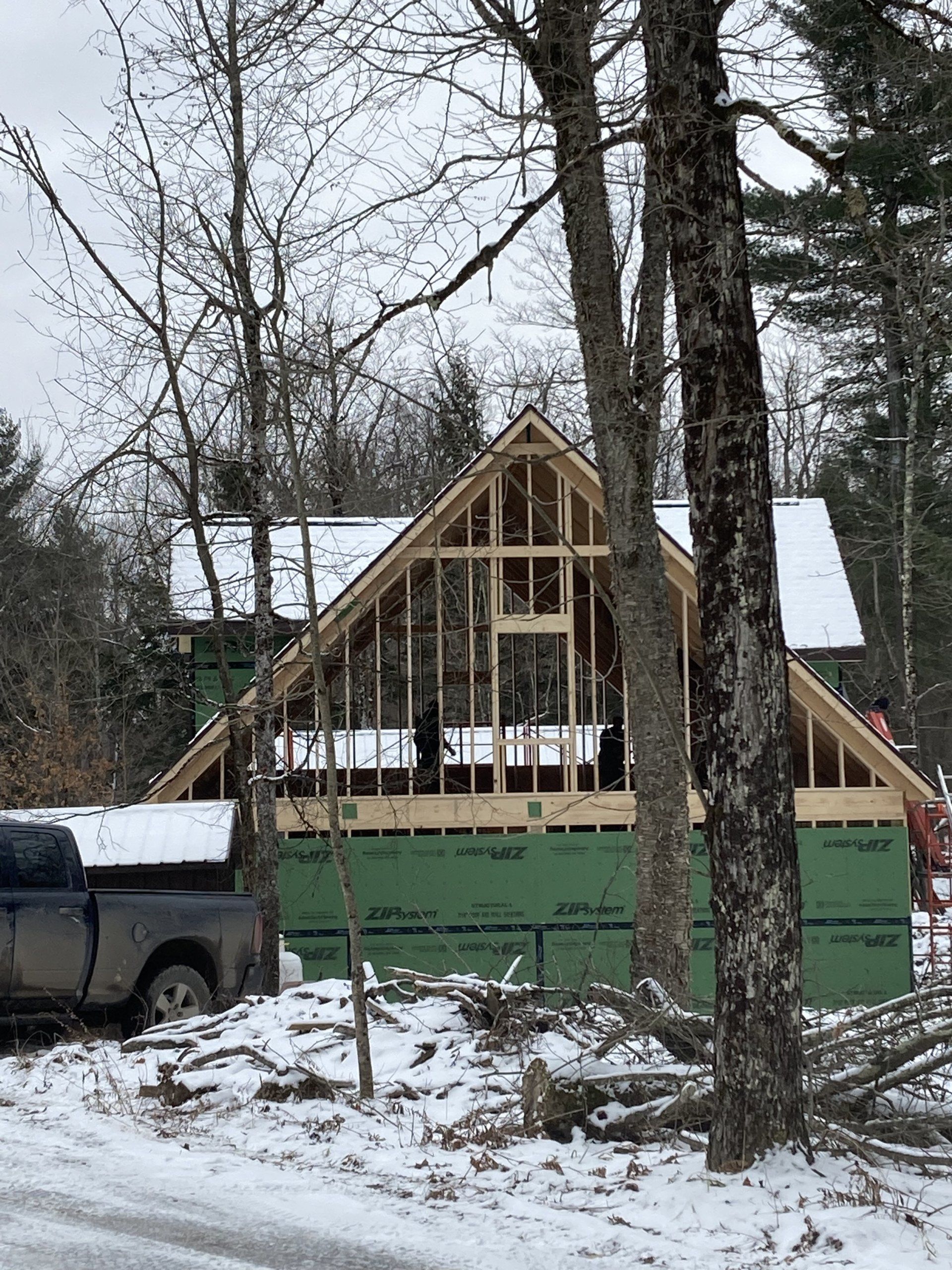 Partially built house with exposed framing and green sheathing in a snowy wooded yard