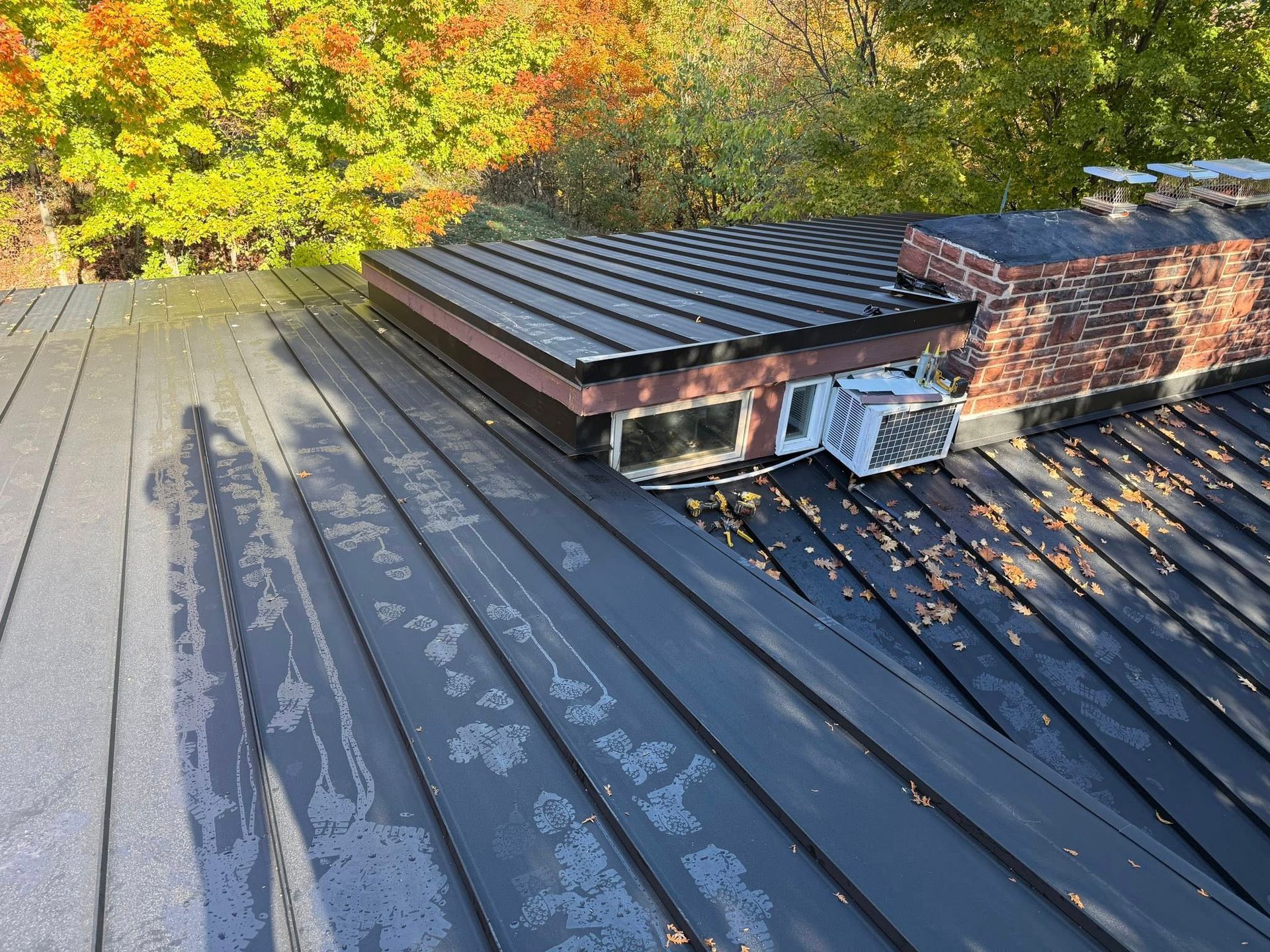 Metal roof with a small brick chimney and fallen leaves near autumn trees