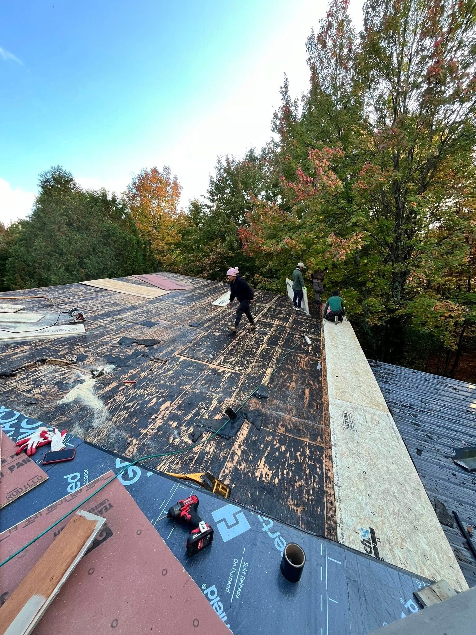 Workers repairing a shingle roof with tools spread across the surface, beside trees and a gravel area.