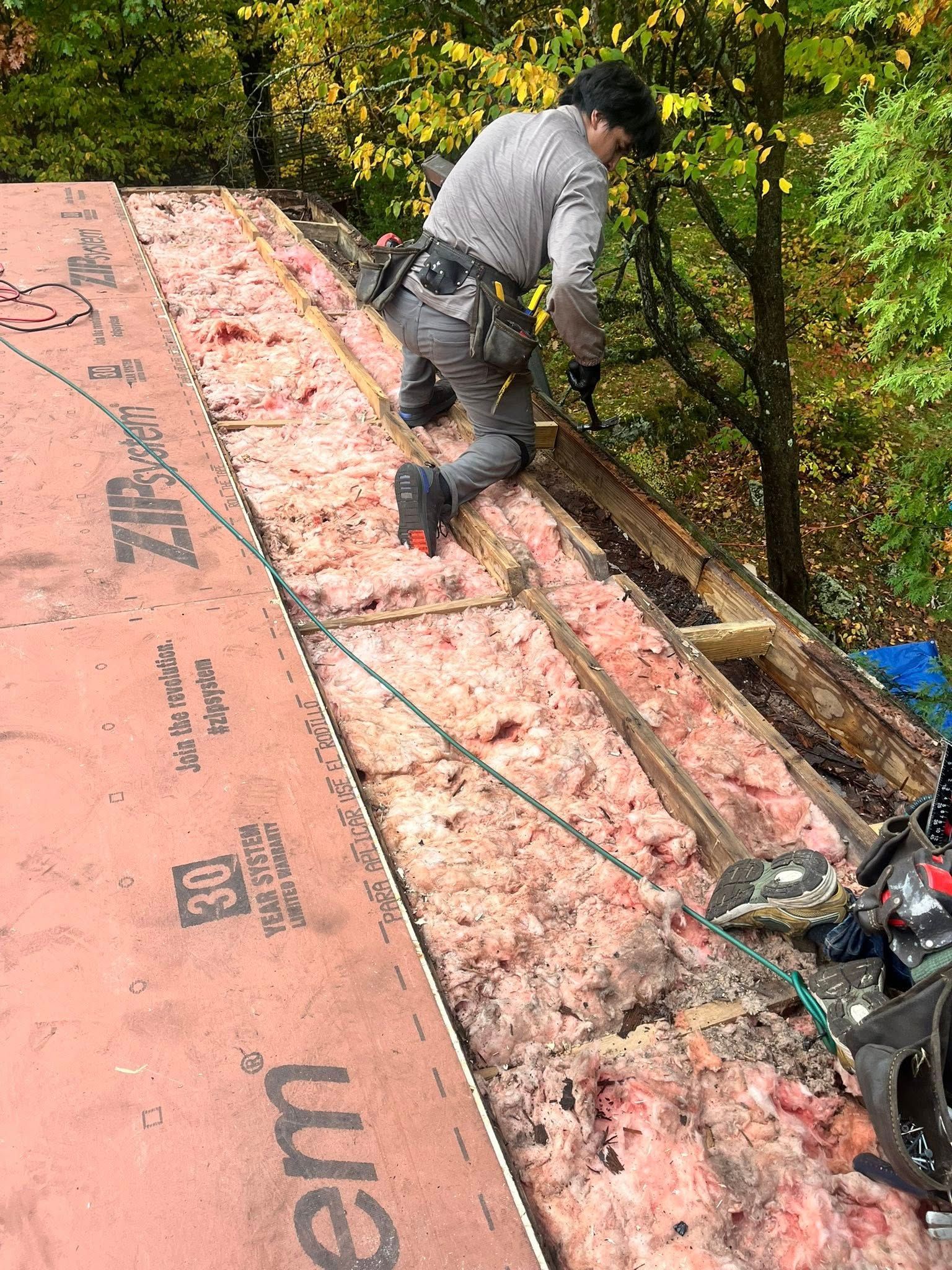 Worker kneeling on a roof installing pink underlayment beside trees