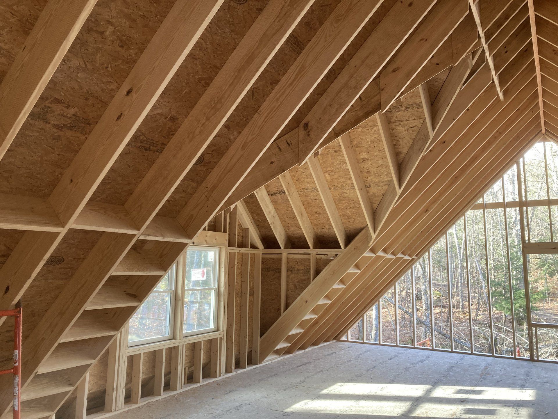 Unfinished wooden house interior with exposed roof trusses and large windows overlooking trees.
