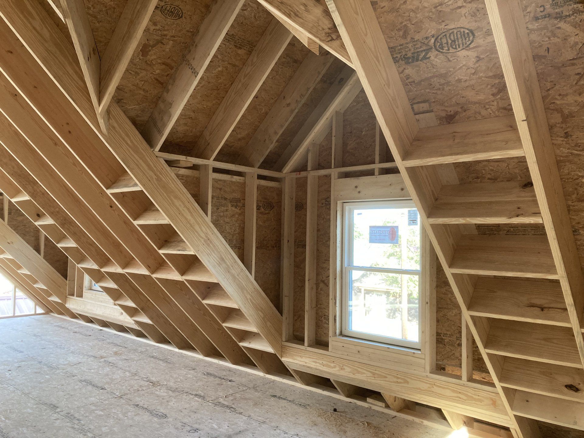 Unfinished attic with exposed wooden rafters, stairs, and a window letting in daylight.