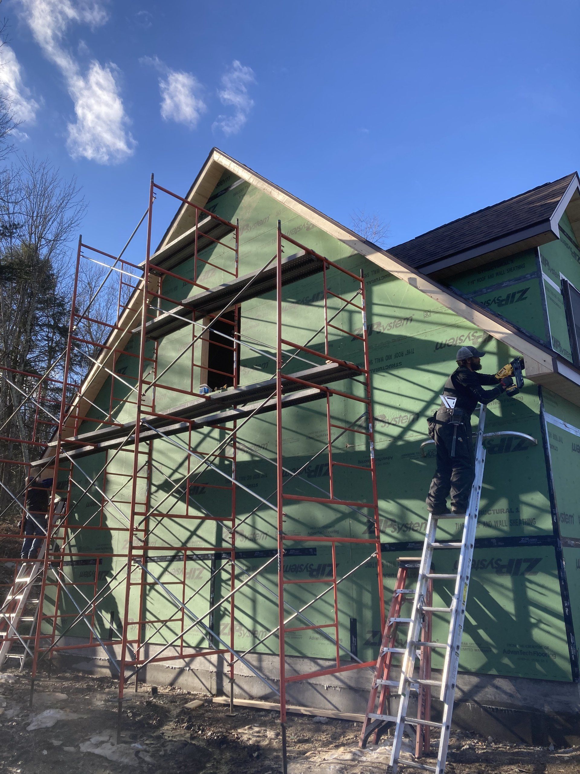 Two workers on scaffolding and a ladder painting a green house exterior under a blue sky