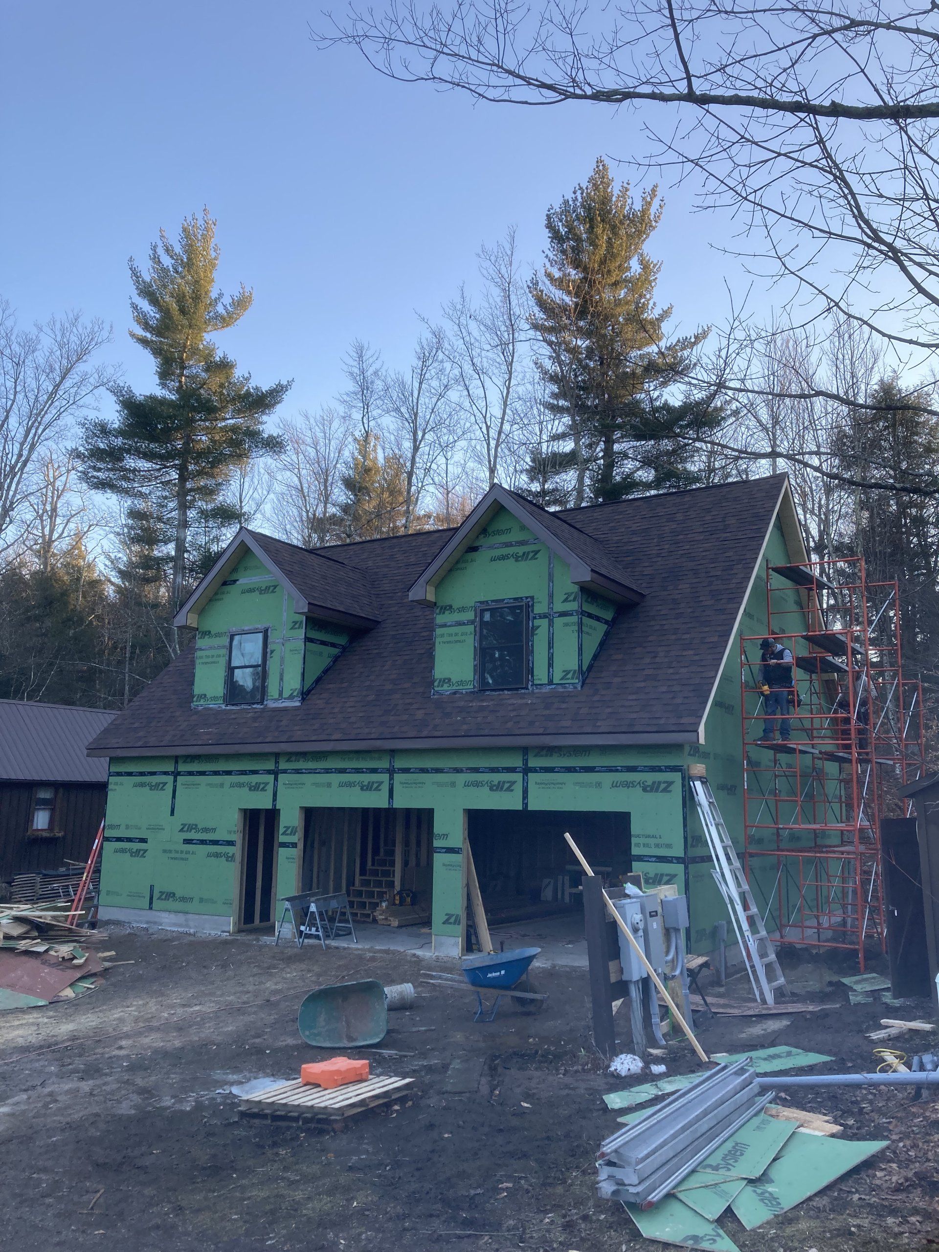 Green house under construction with exposed framing, ladders, and building materials in the yard.