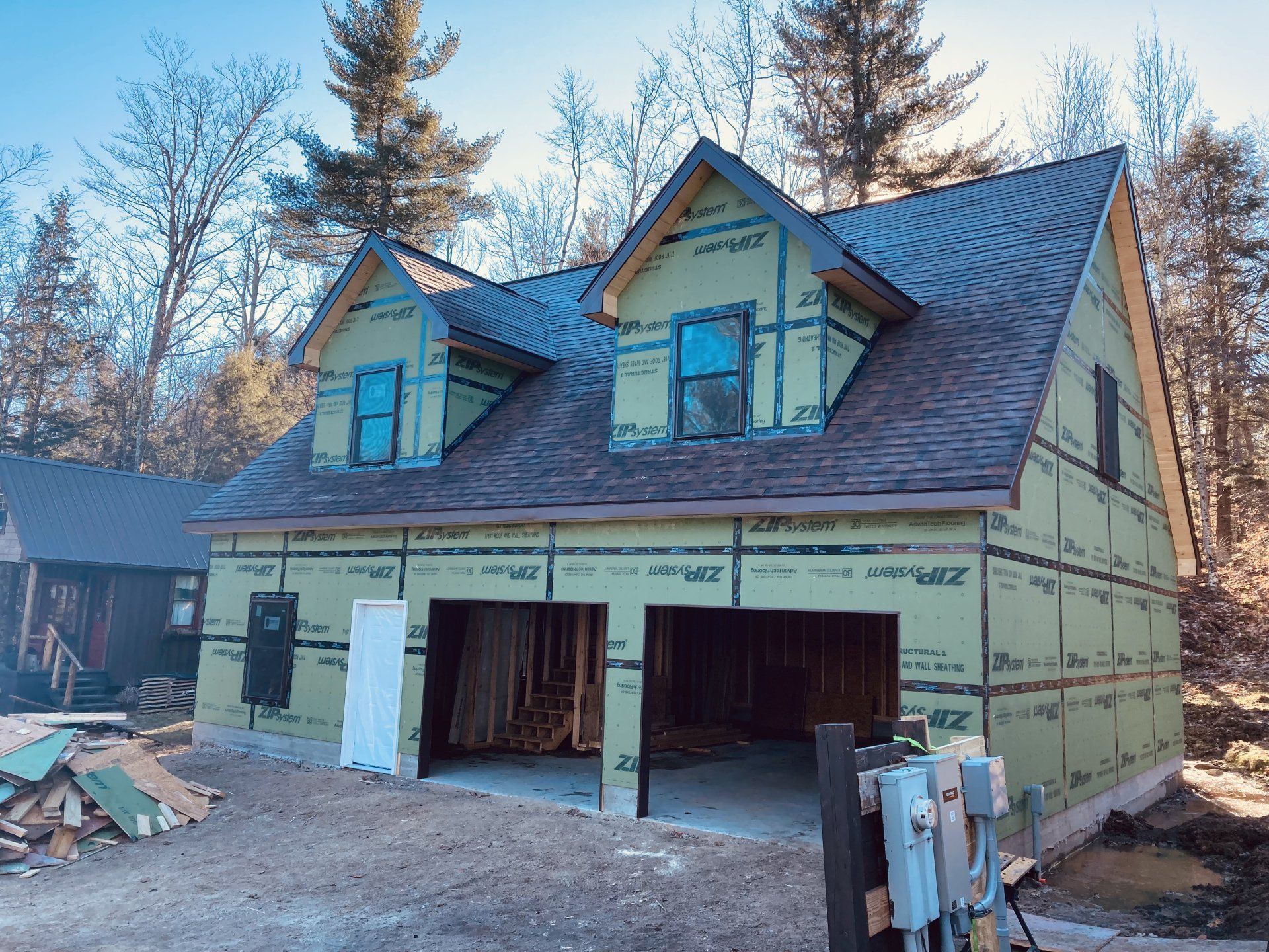 Partially built house with green wrap, open garage, and piles of lumber in a wooded yard