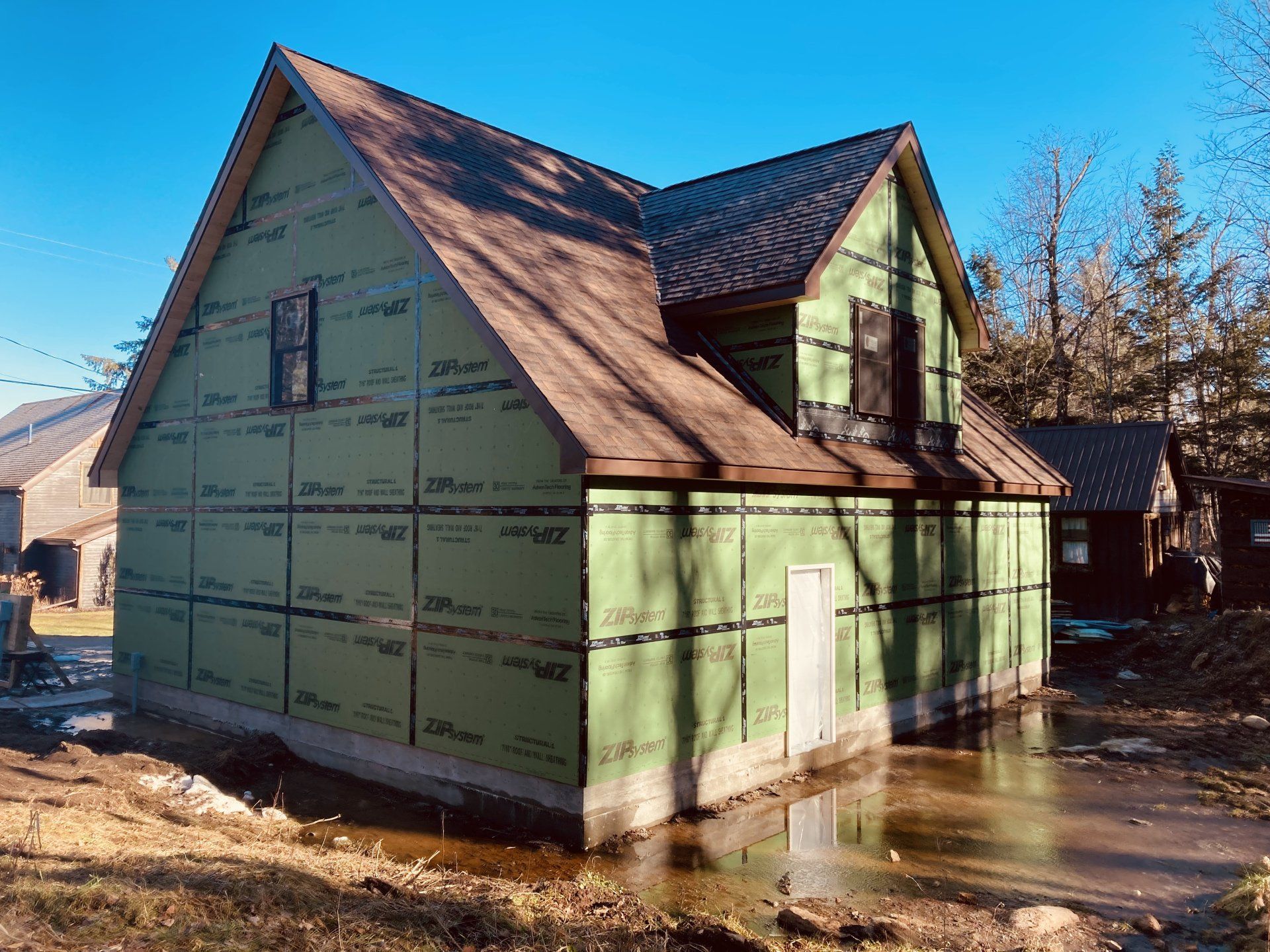 Unfinished house with green exterior wrap and steep roof in a muddy construction site under blue sky