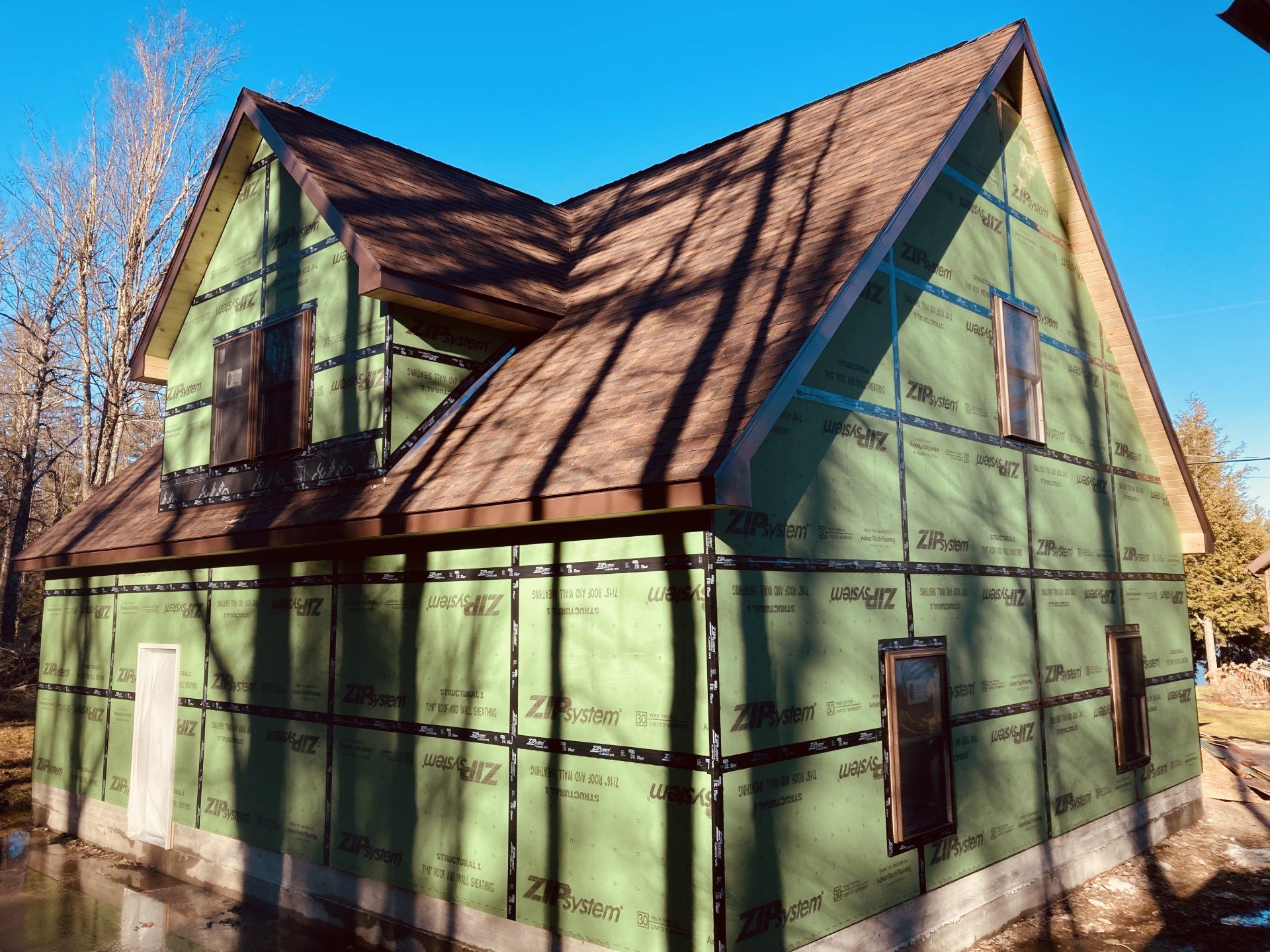 Green house under construction with exposed framing, wrap, and steep roof in a sunny yard