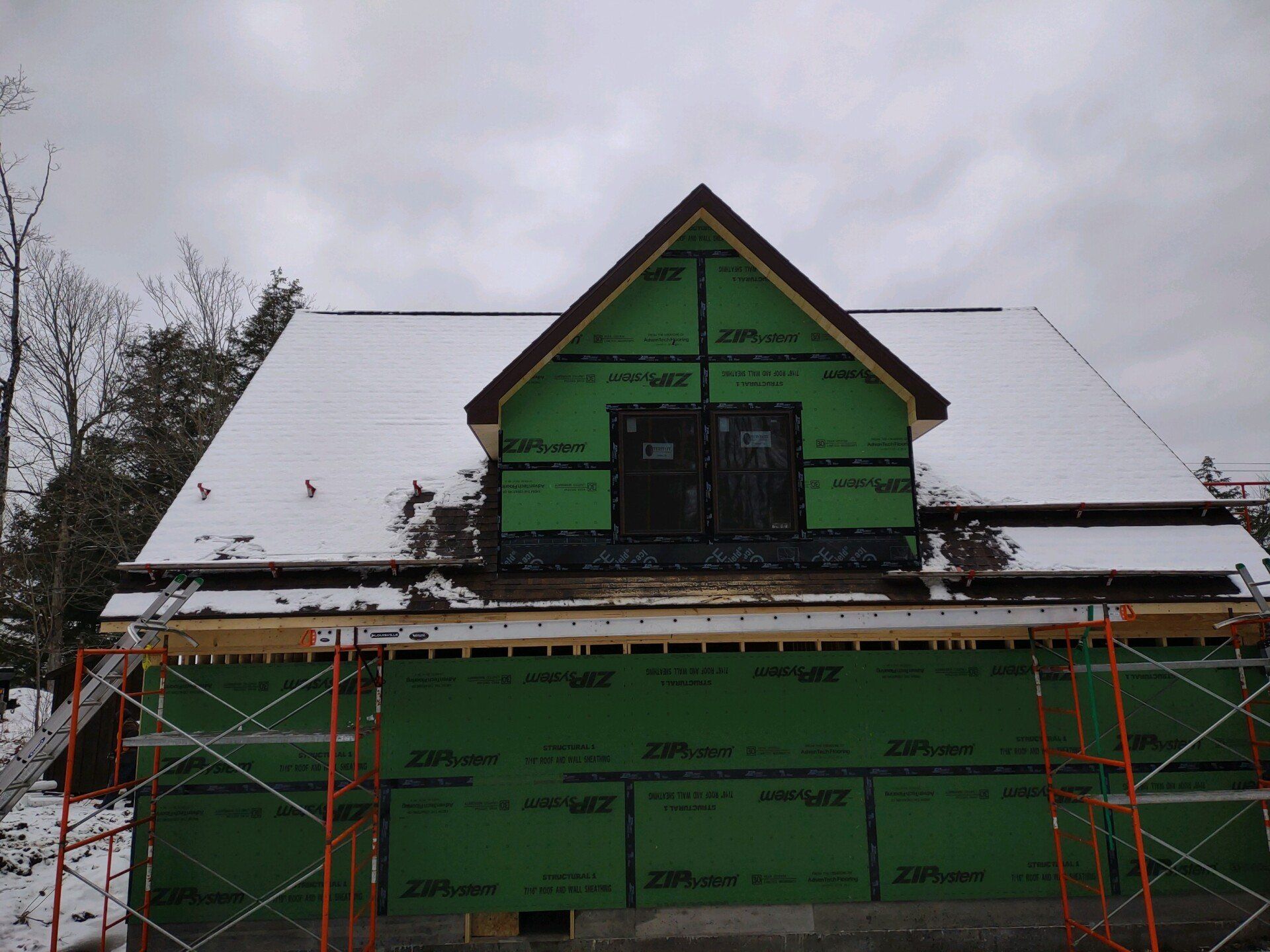 Partially renovated house with green insulation wrap and snow-covered roof under overcast sky