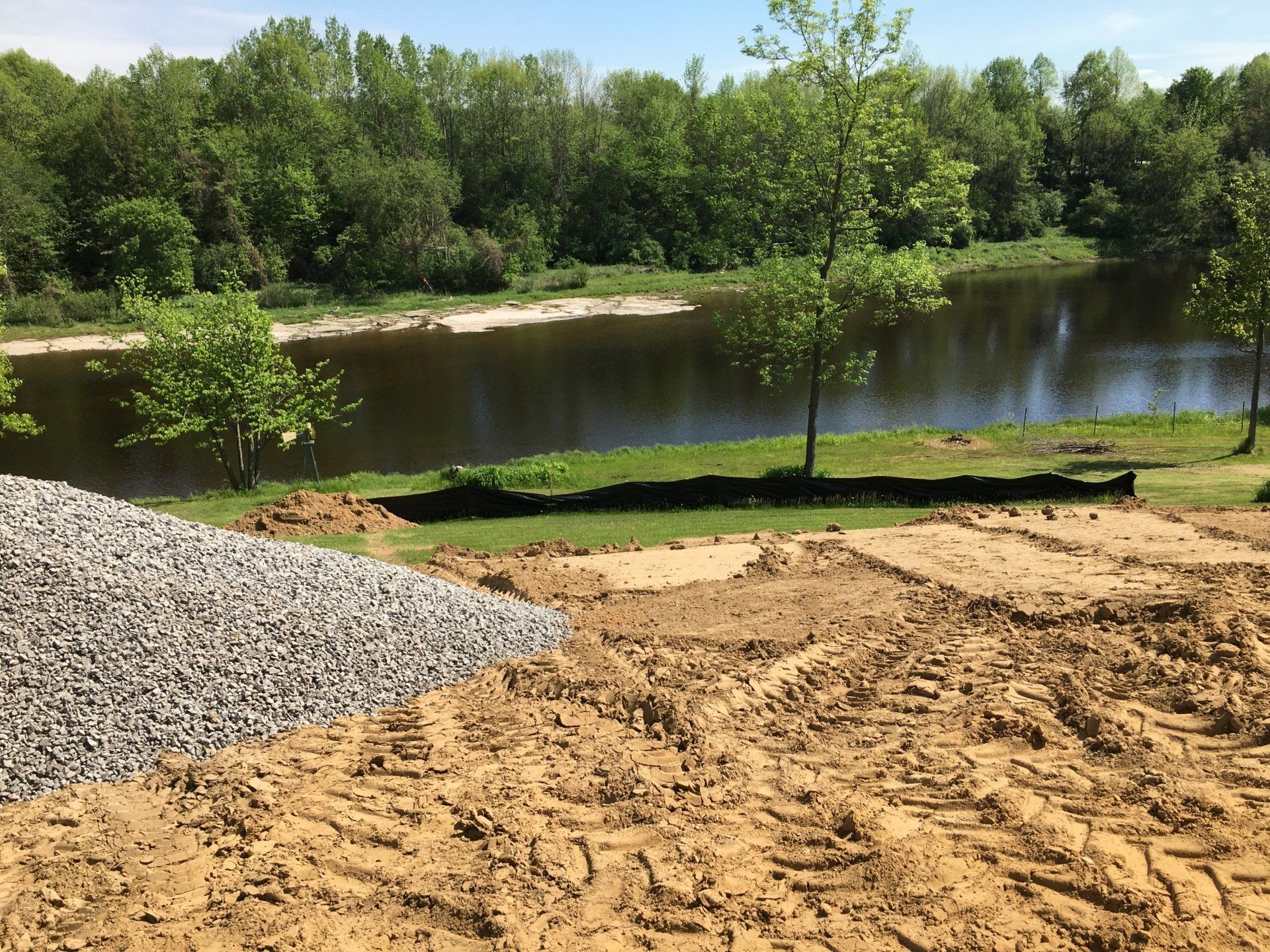 Riverbank with fresh sand, gravel, and a grassy edge beside calm water and trees