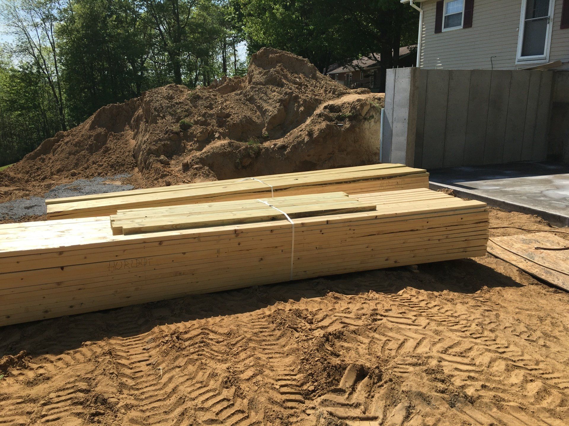 Long lumber beams stacked on a dirt construction site beside a house and fence.