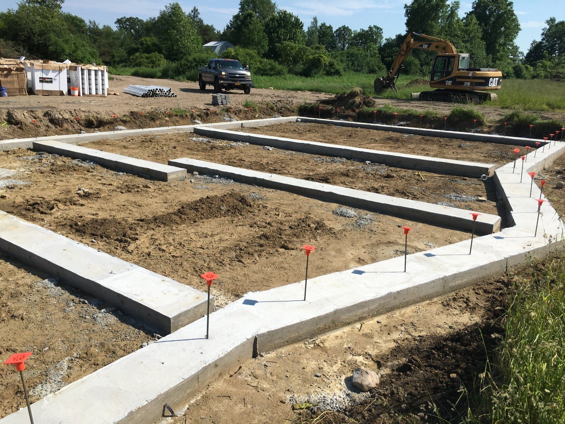 Concrete foundation footings laid out on a construction site with dirt, stakes, and excavator in the background