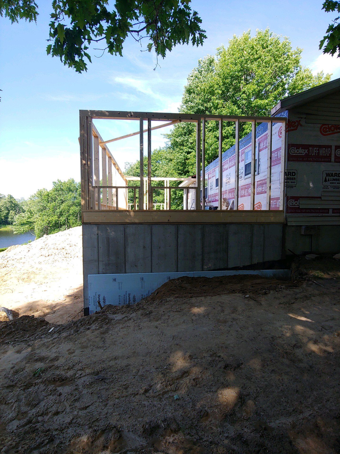 Partially built wooden deck beside a river, with trees and bright sky in the background