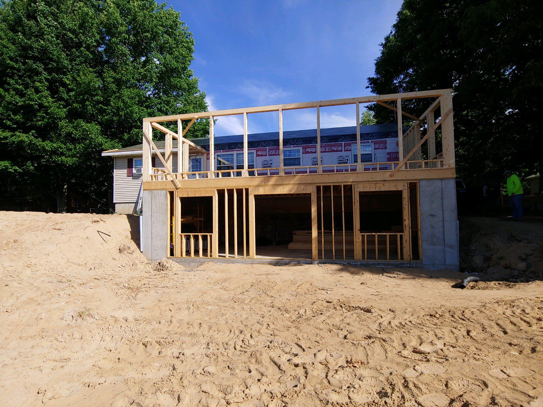 Partially built house with exposed framing on a sandy lot surrounded by trees