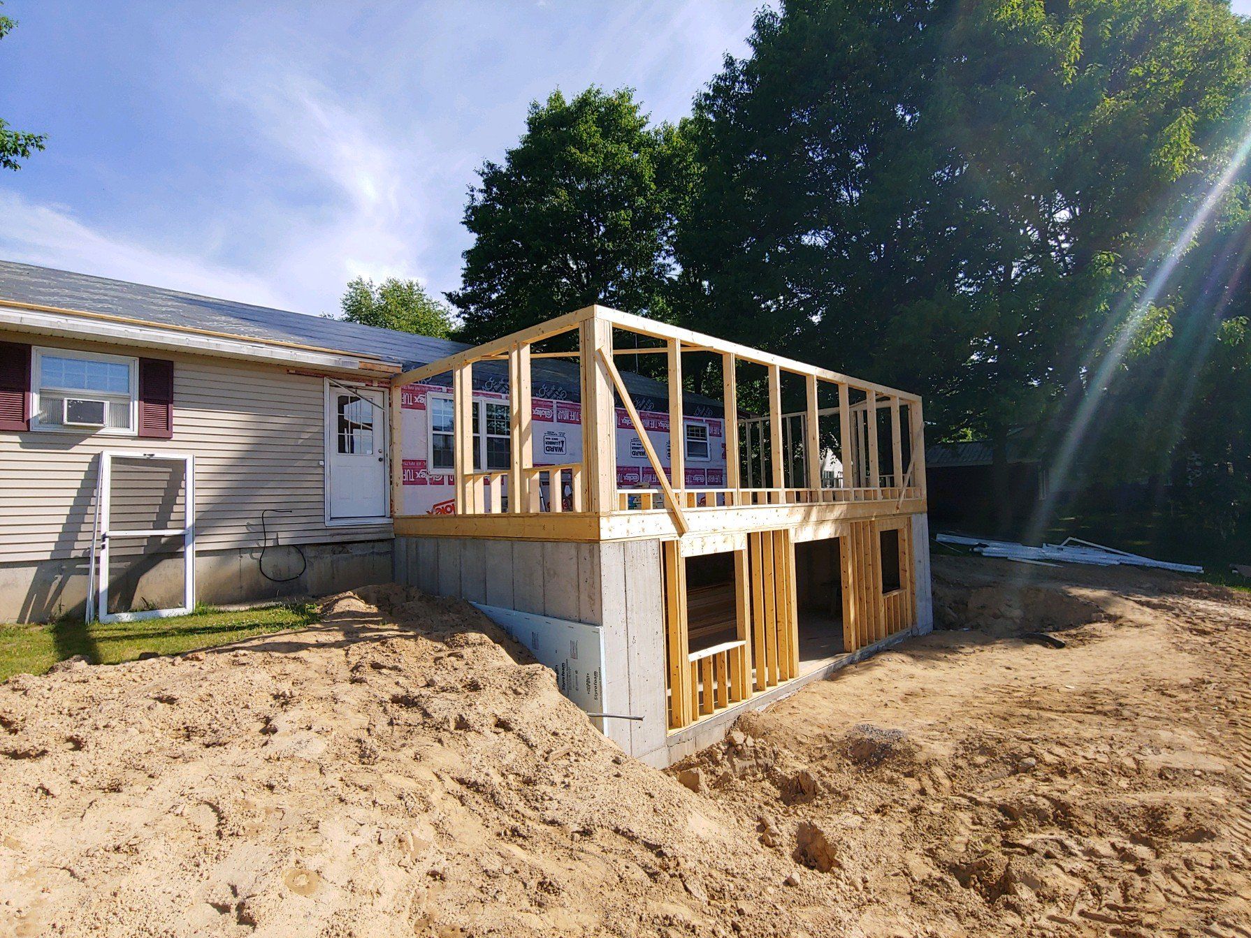 Partially built wooden deck extension beside a house, with exposed framing and sandy ground around it