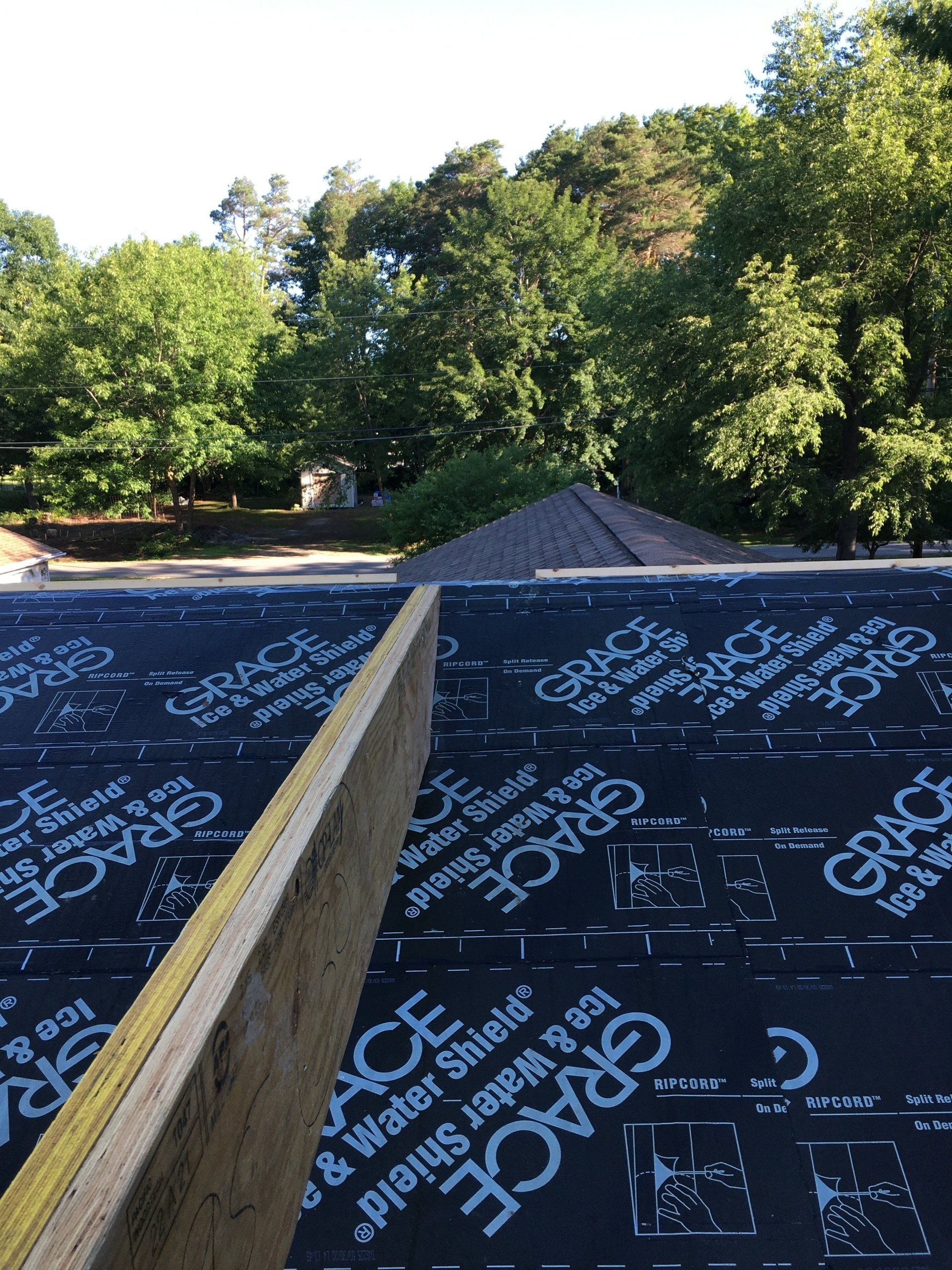 Roofer installing black underlayment on a pitched roof with blue lettering, surrounded by green trees
