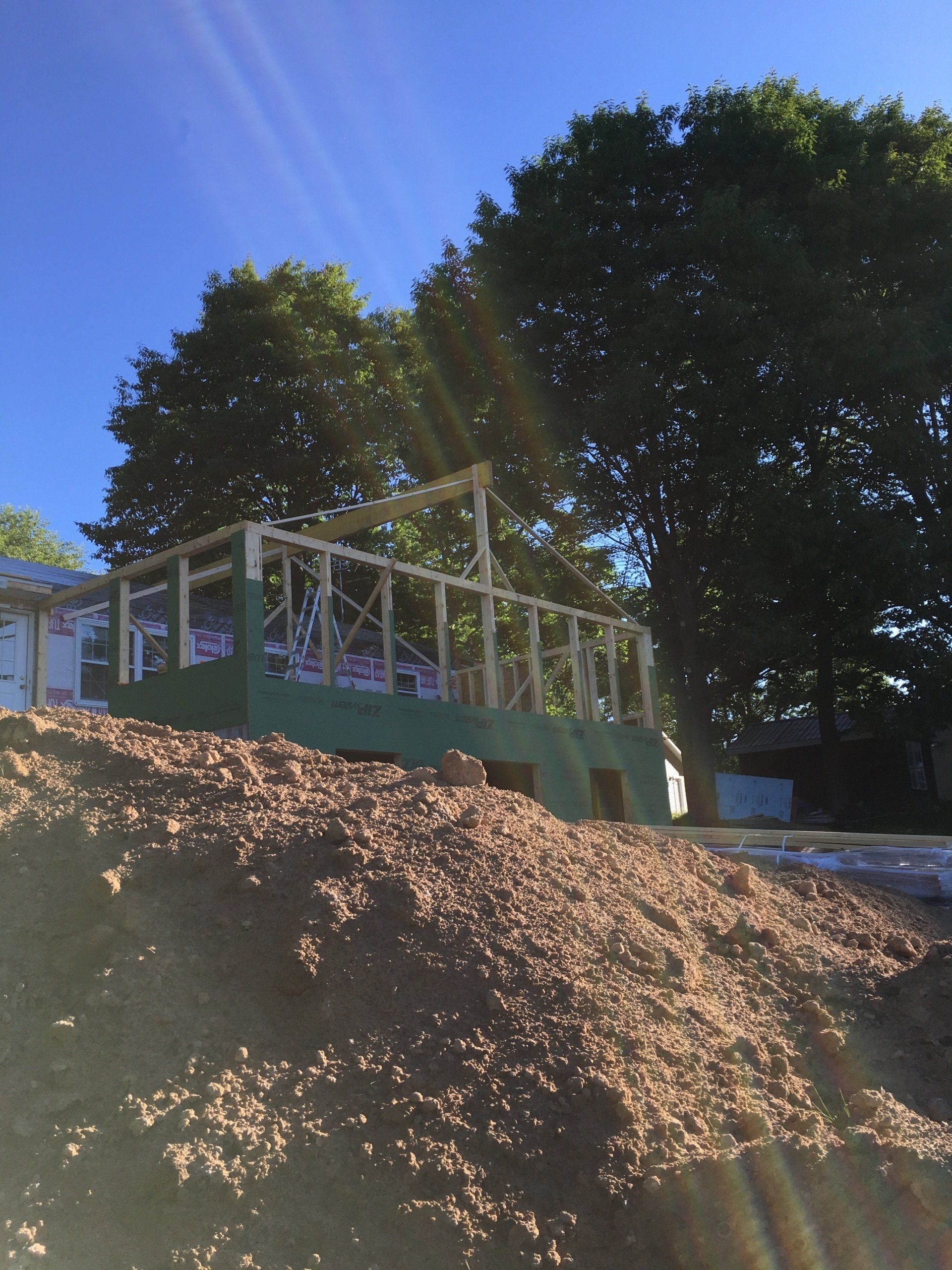 Raised deck under construction on a hillside, with lumber framing and large trees against a blue sky.