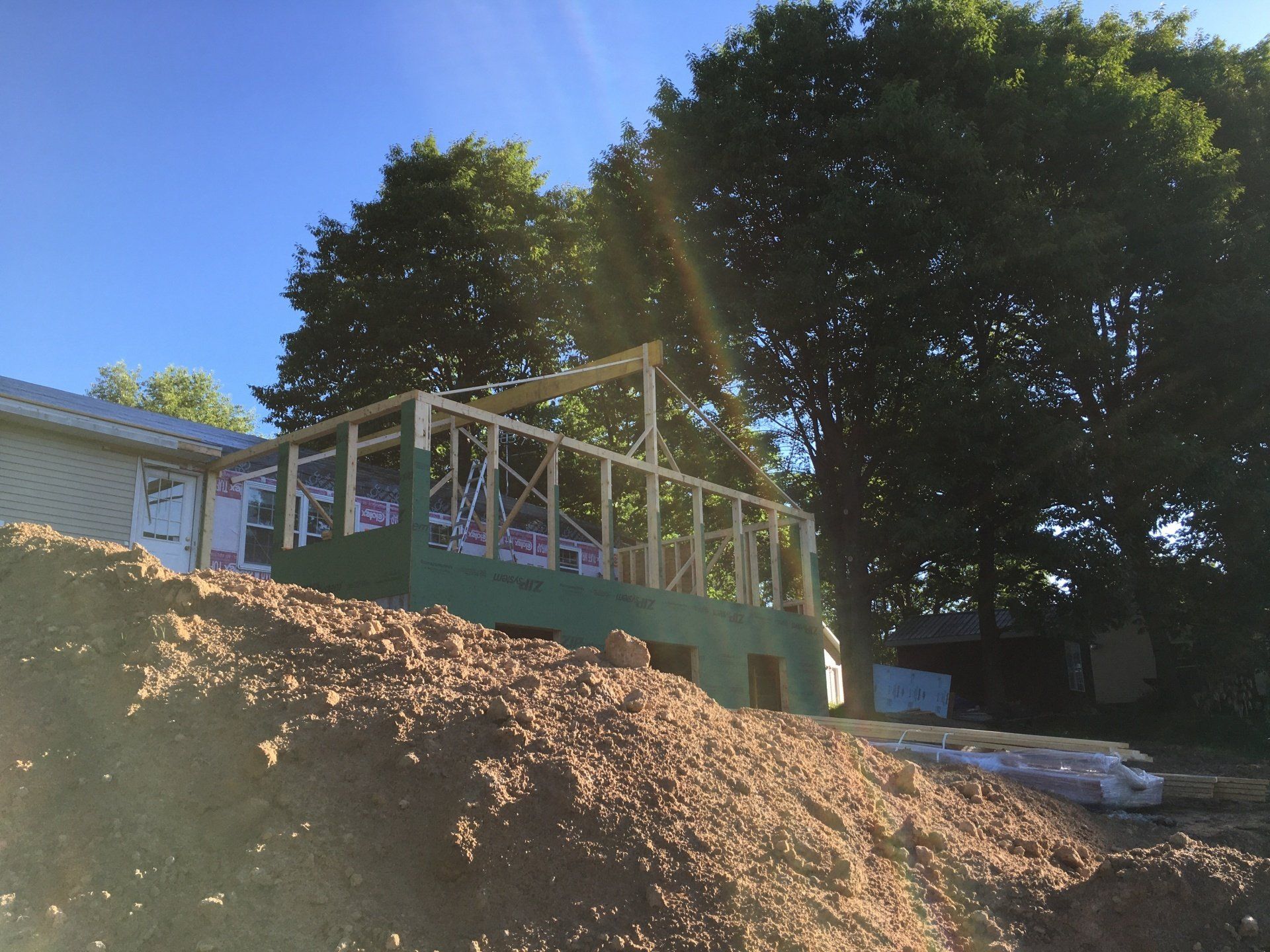 House under construction beside a dirt slope, with wooden framing and tall trees in the background