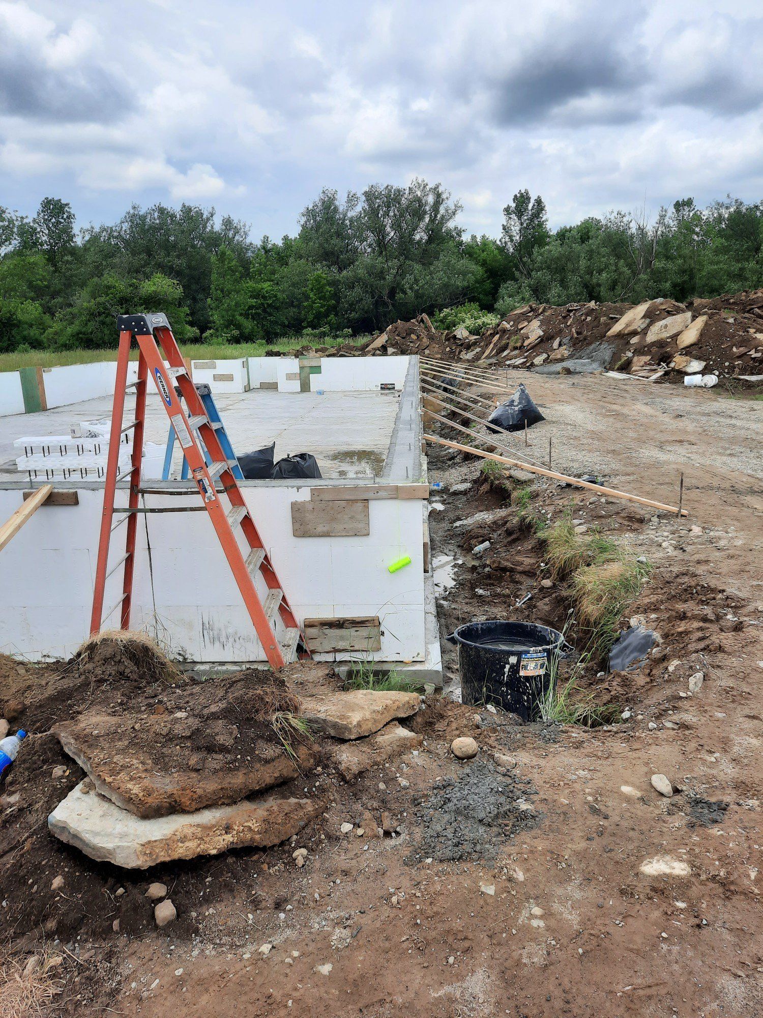 Construction site with foundation walls, orange ladder, and dirt excavation under a cloudy sky