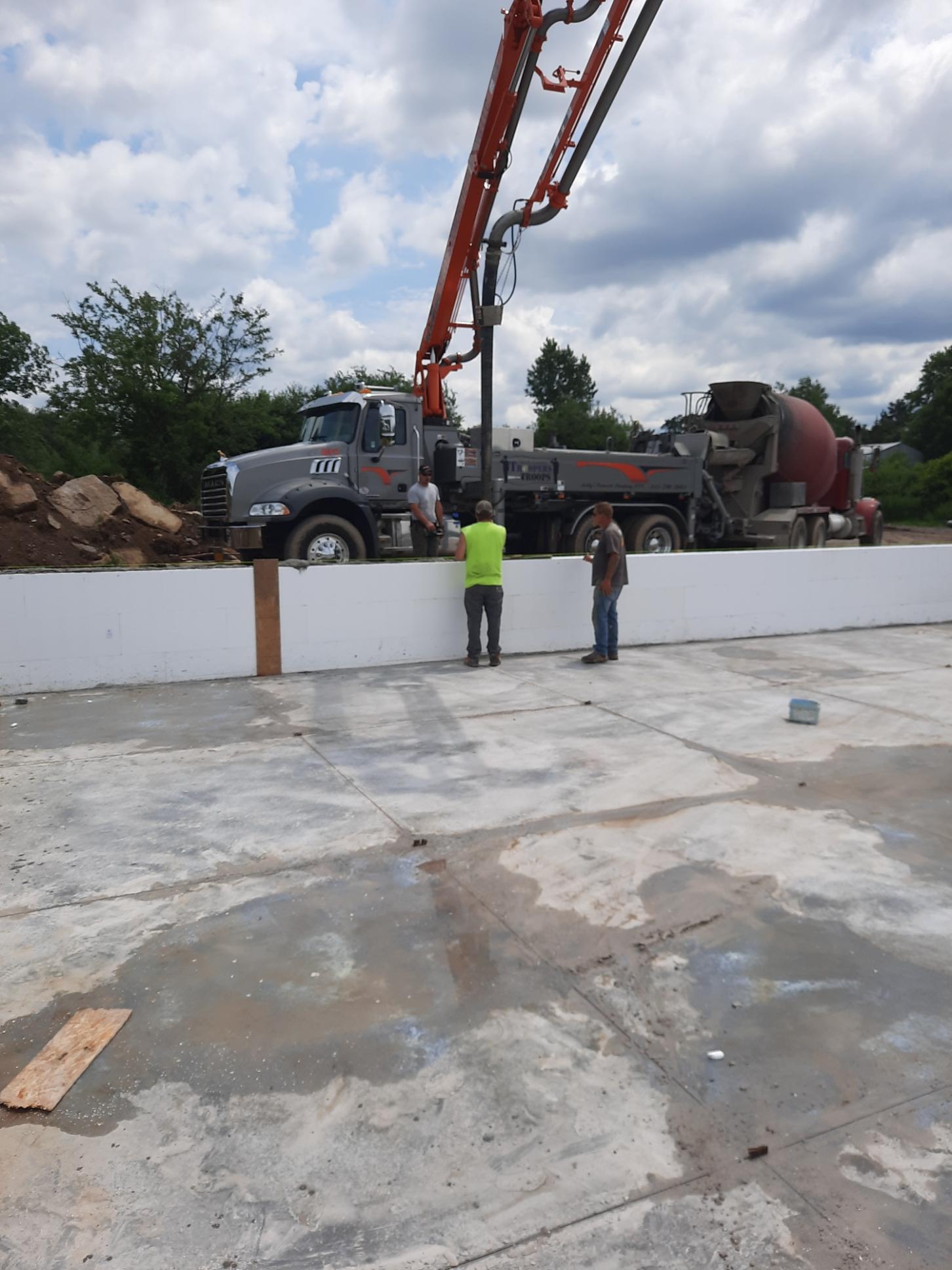 Concrete truck and workers at a construction site, with a pump boom over a slab under cloudy skies