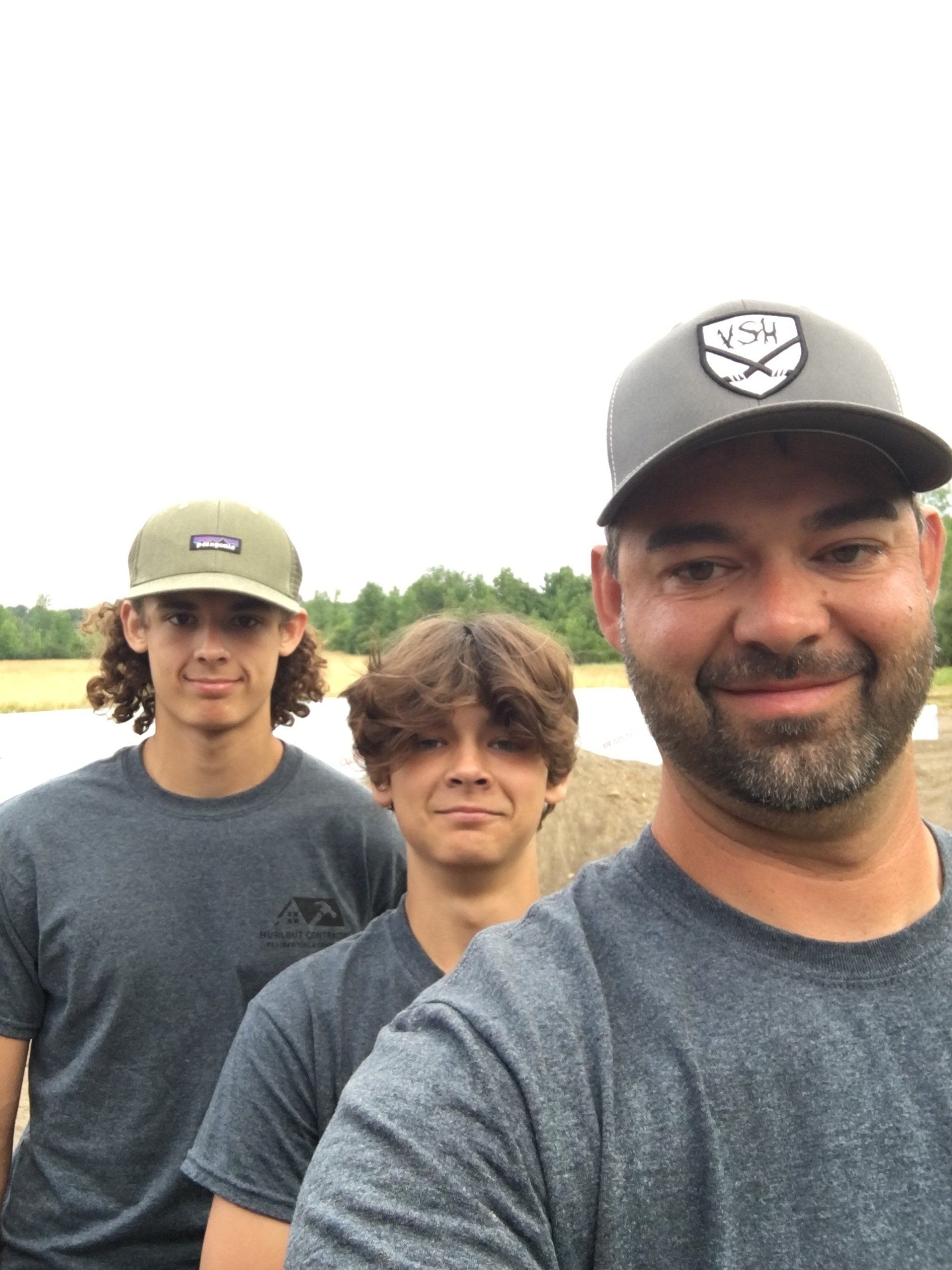 Brian and his sons taking a selfie outdoors, wearing gray shirts and caps, with trees in the background