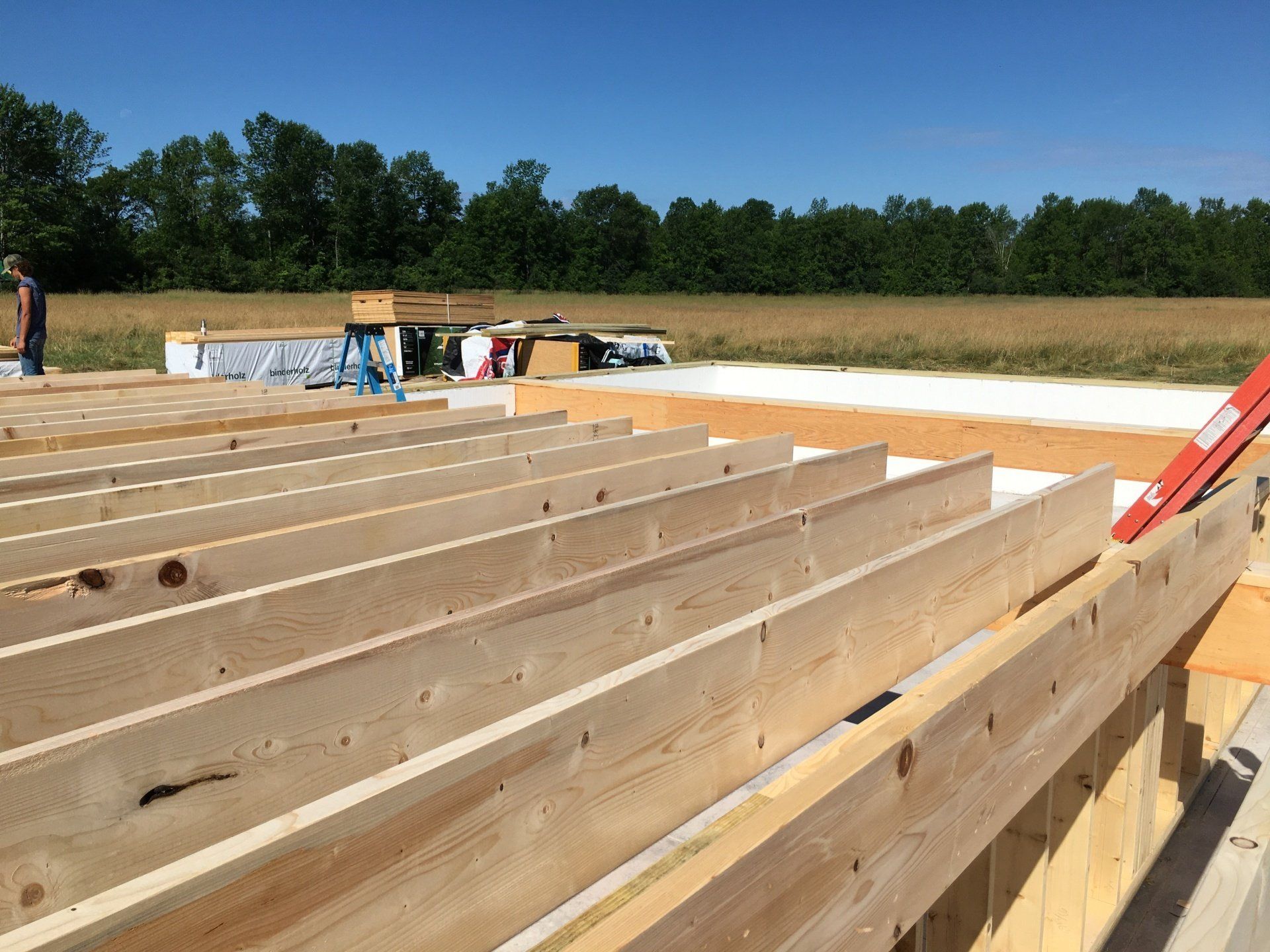 Wood framing under construction at a building site, with workers in the background and a tree line beyond.