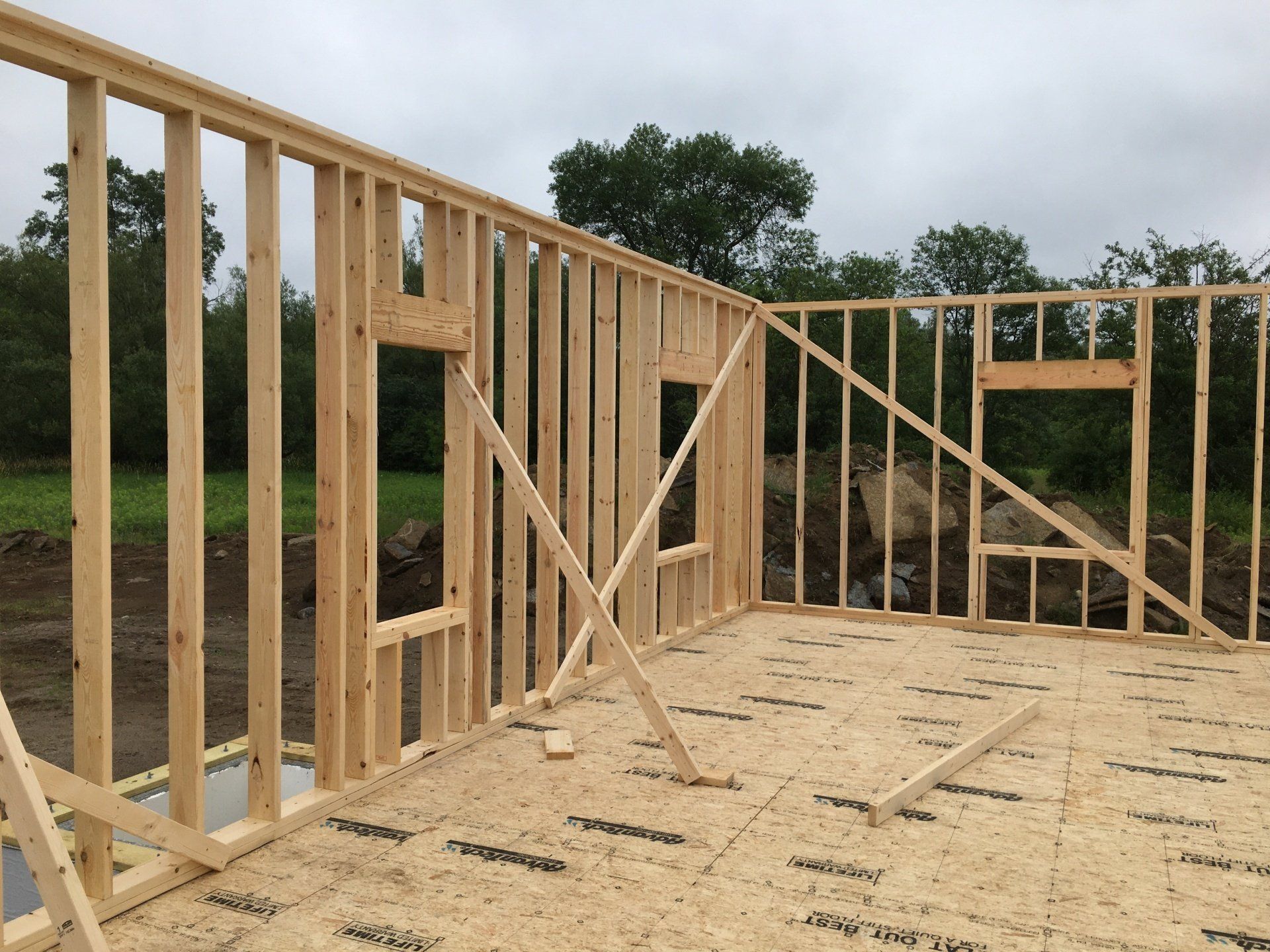 Wooden house frame under construction on a plywood floor, with exterior walls and roof framing visible.