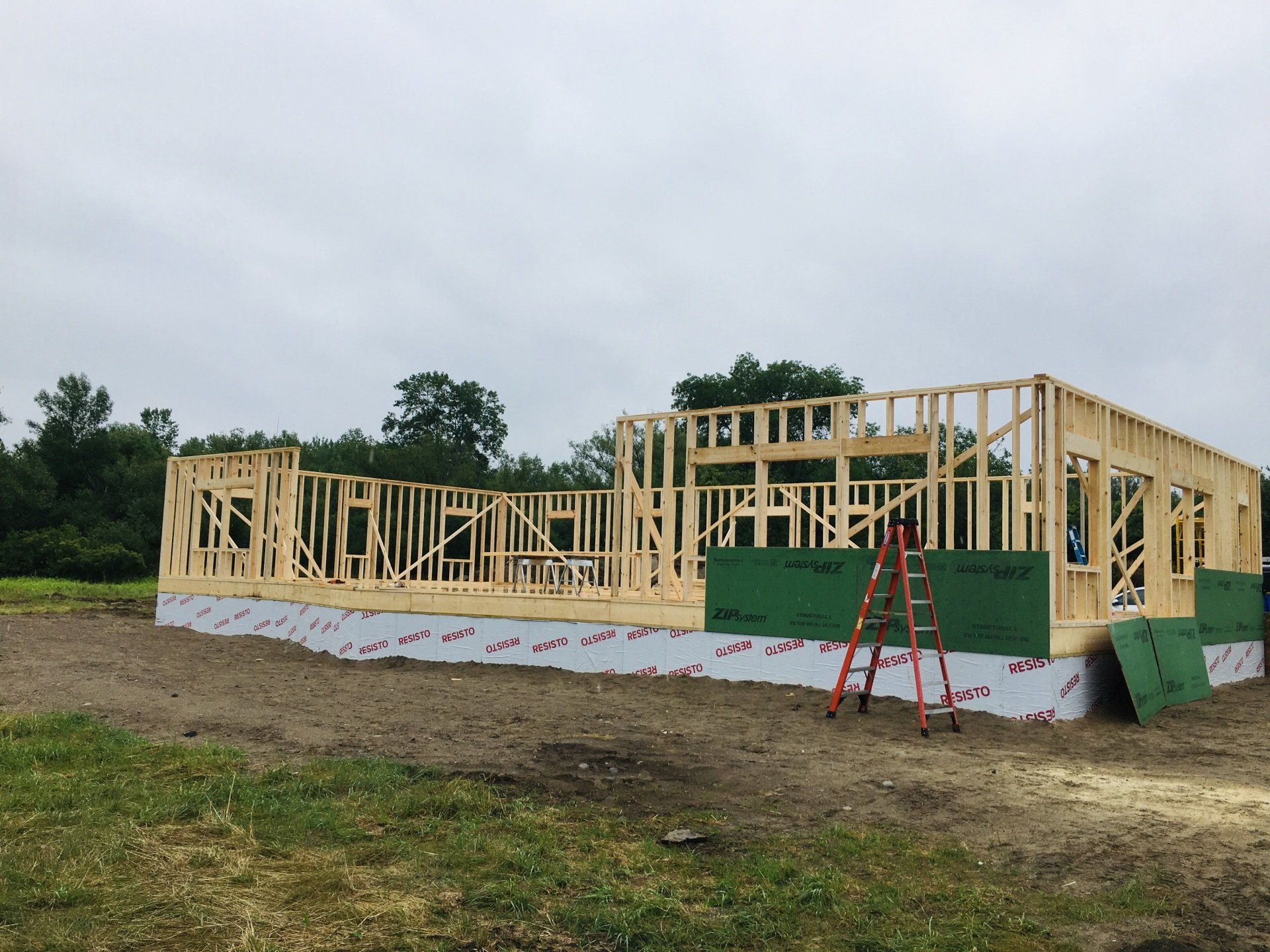 Wood-framed house under construction on a grassy lot, with a ladder and green sheathing panels.
