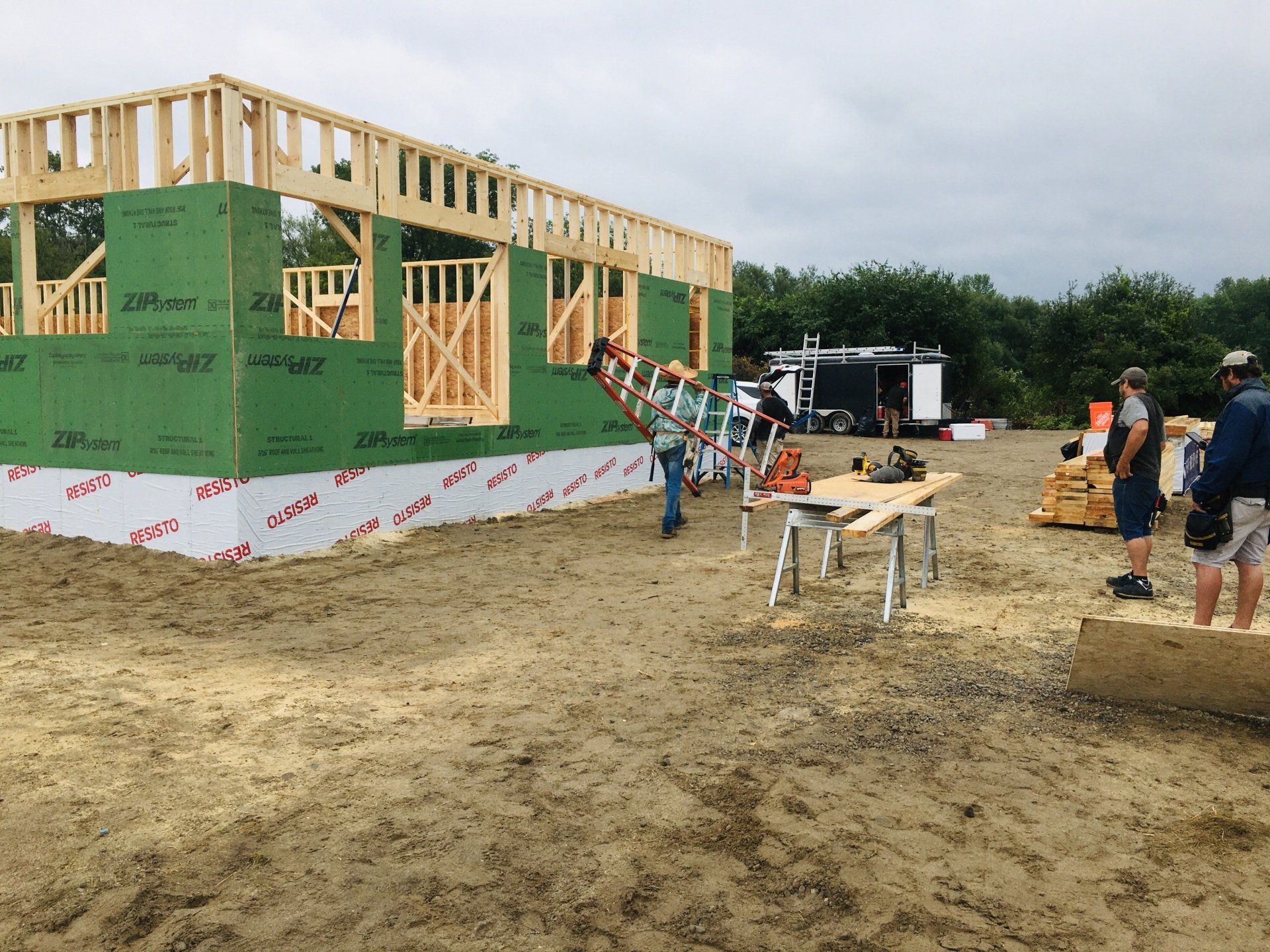 Construction workers framing a green house on a muddy lot with lumber, tools, and a truck nearby