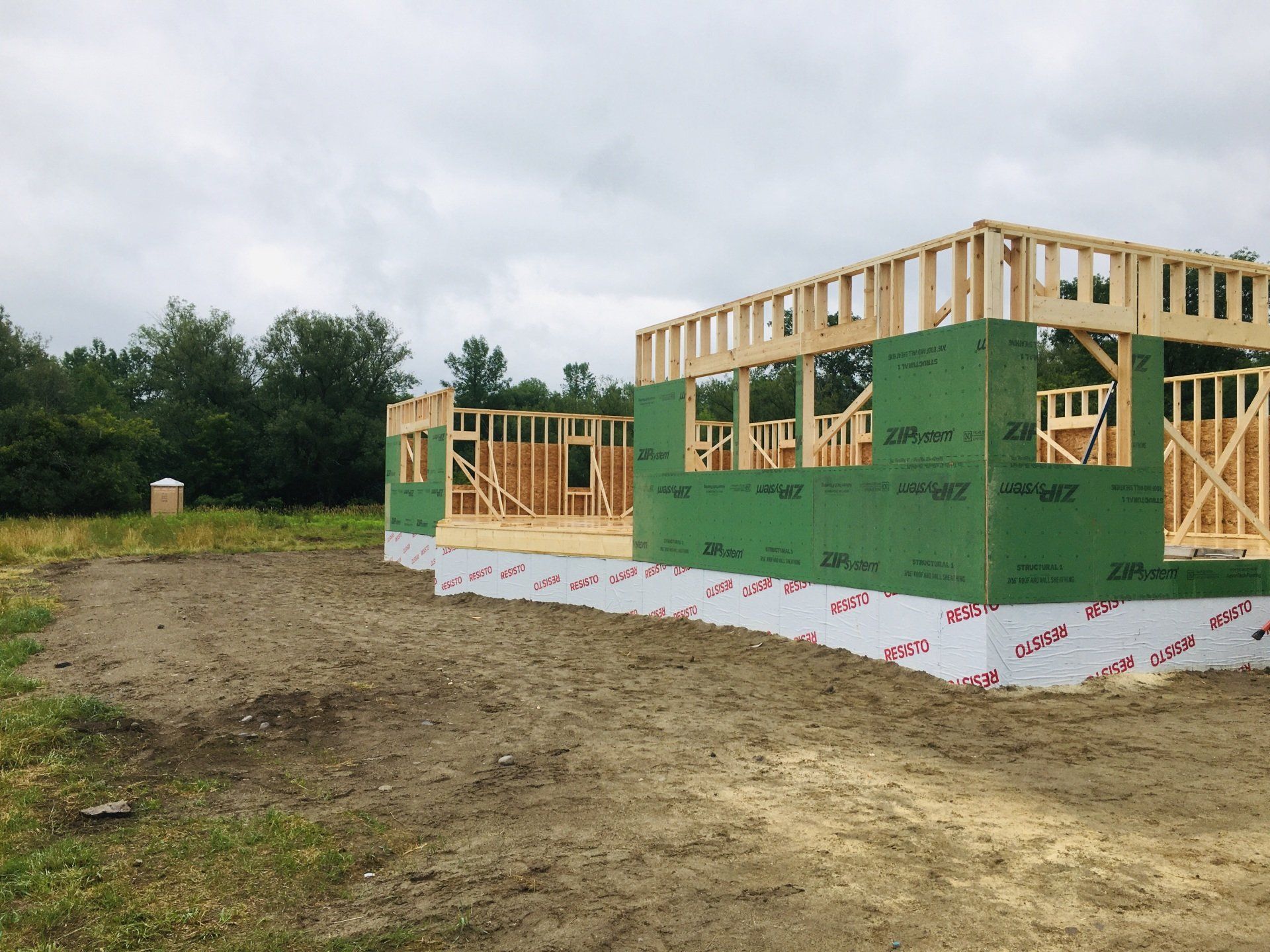 Partially built house frame with green sheathing on a dirt lot under a cloudy sky