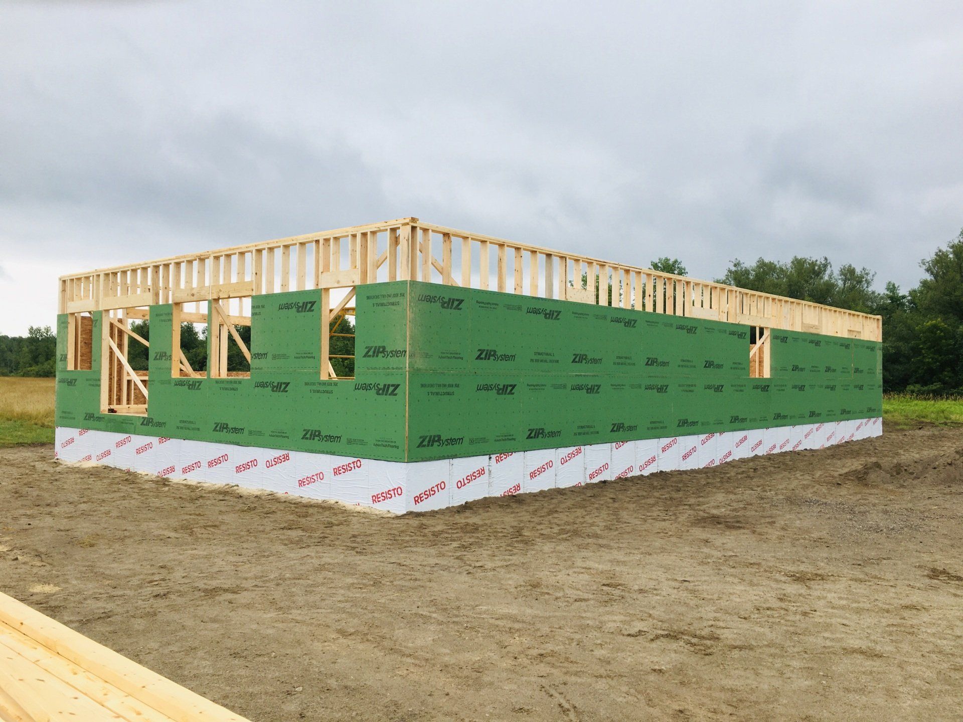 Partially built house frame wrapped in green sheathing on a dirt lot under cloudy sky