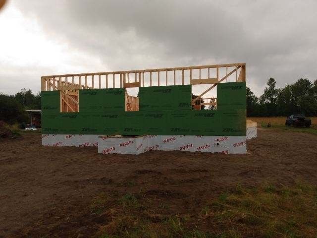 Partially framed house with green house wrap on a muddy lot under a cloudy sky